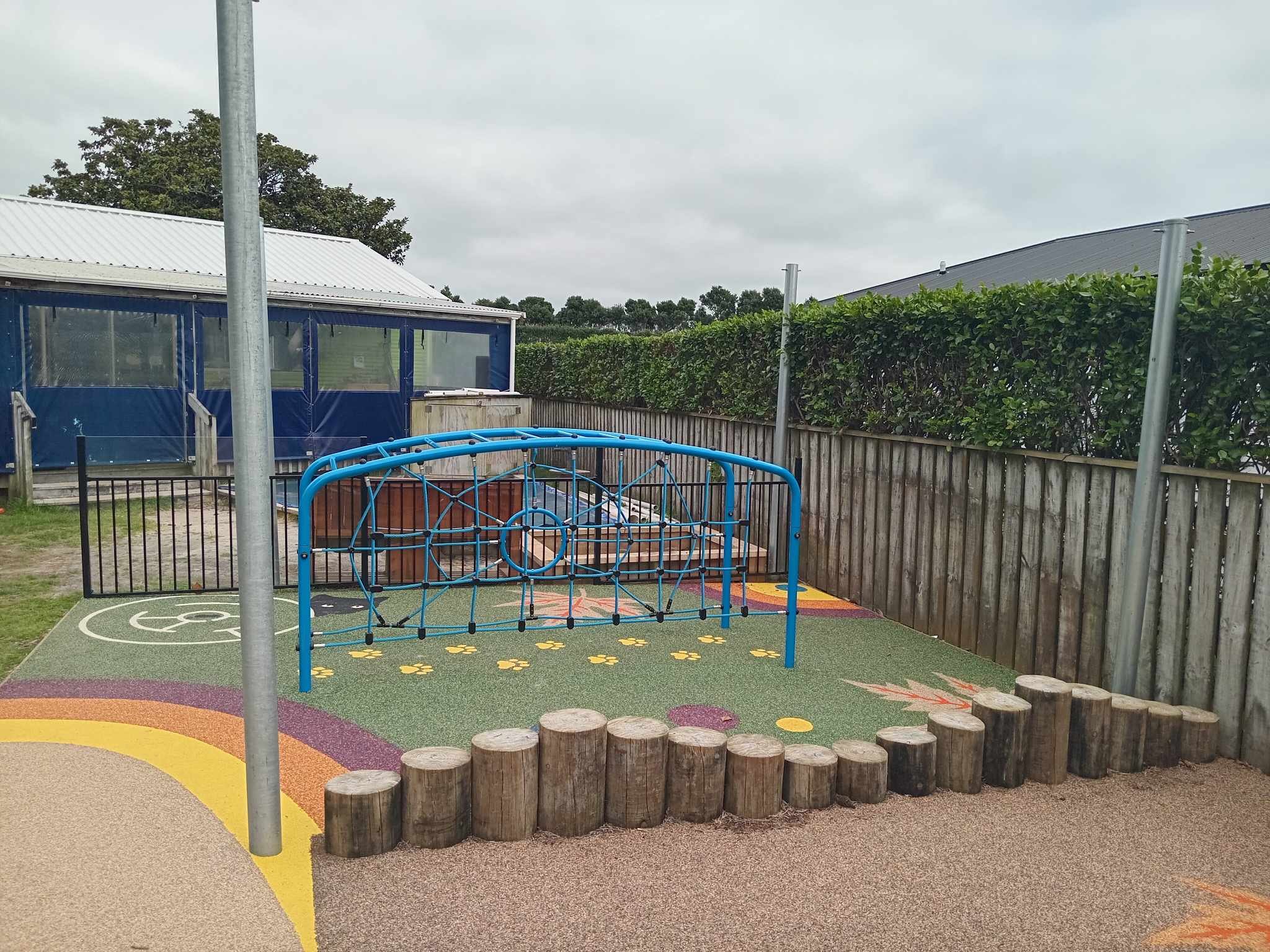 Playground area with blue climbing structure and colorful rubber surface, surrounded by wooden fence and small wooden logs. Nearby building with blue tarp sides.
