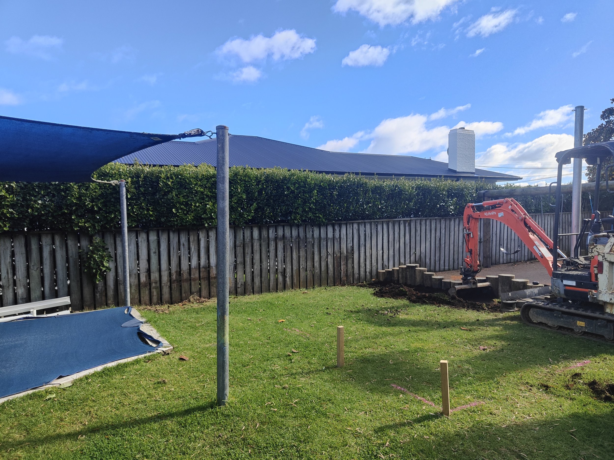 Backyard under construction with excavator, wooden stakes, and partial shade structure.
