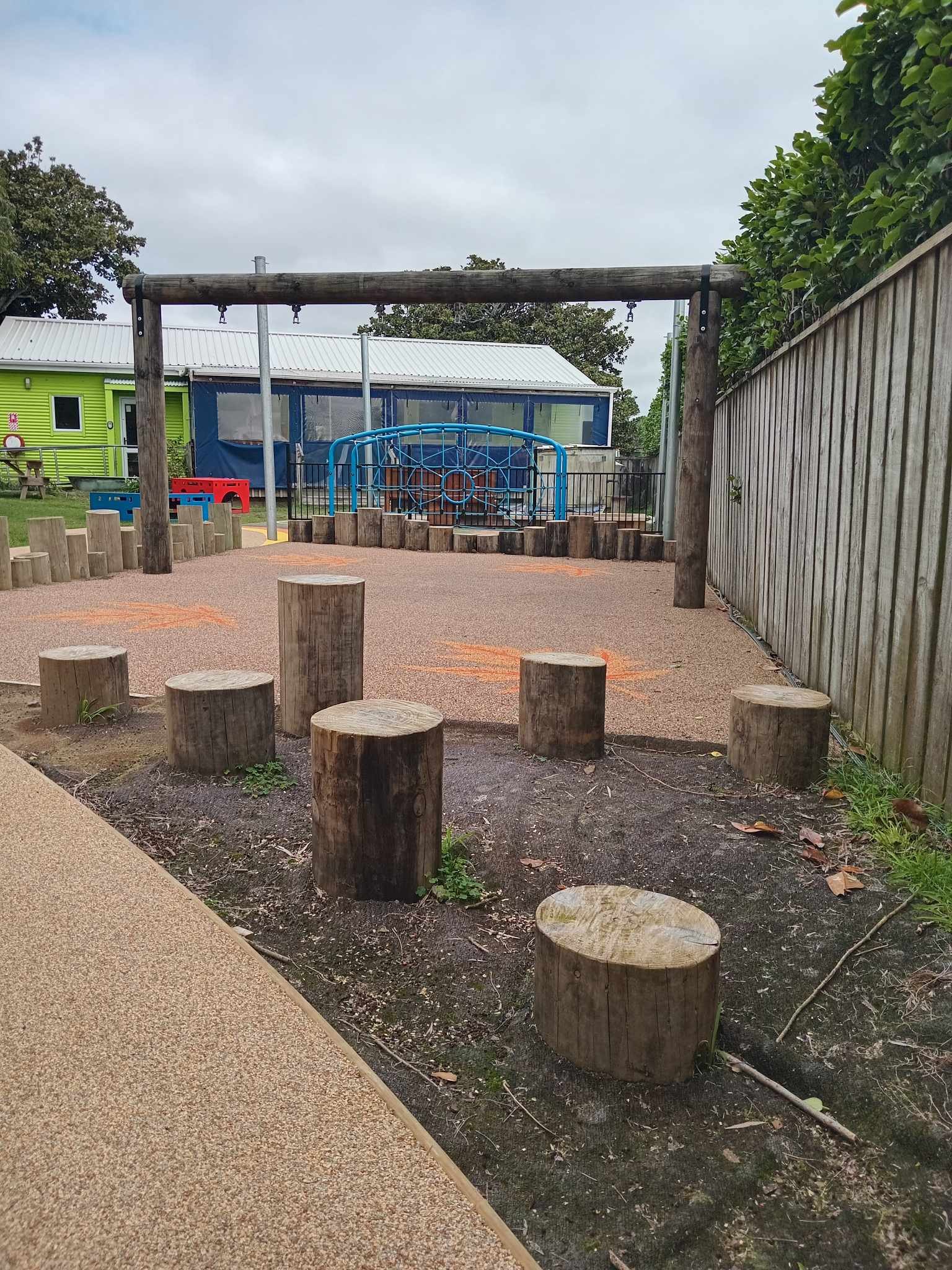 Outdoor playground area with wooden logs, climbing structure, building in background, and surrounding fence.
