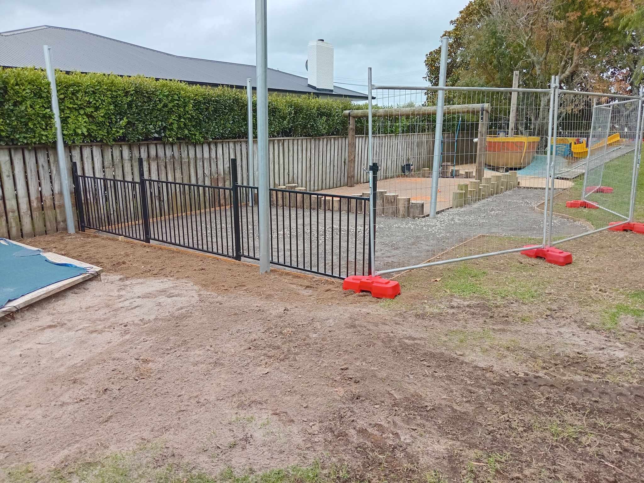 Outdoor construction area with metal fencing and a wooden barrier. Grounds partially covered with dirt and a tarp. Some playground equipment visible in the background.