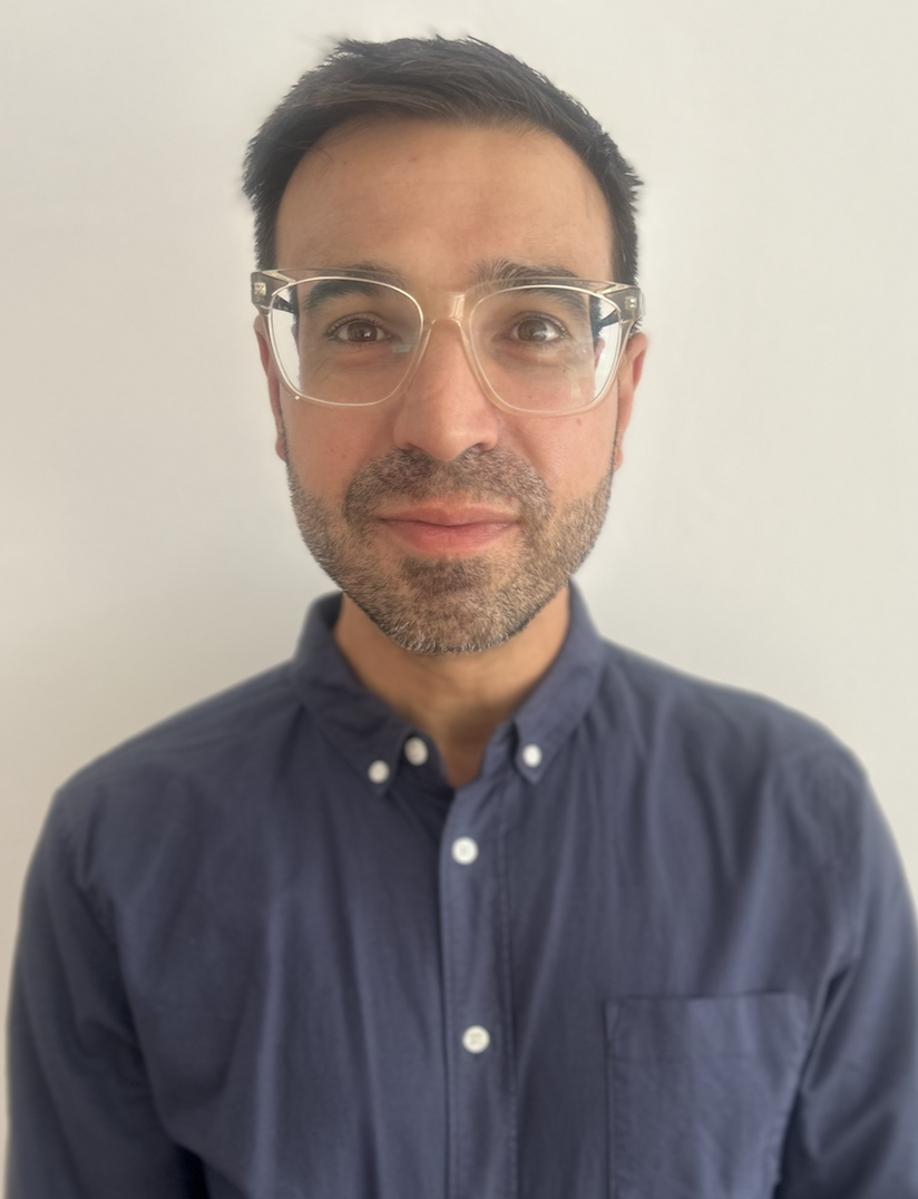 A man with glasses, short dark hair, and a beard wearing a navy blue button-up shirt standing against a plain light-colored background.