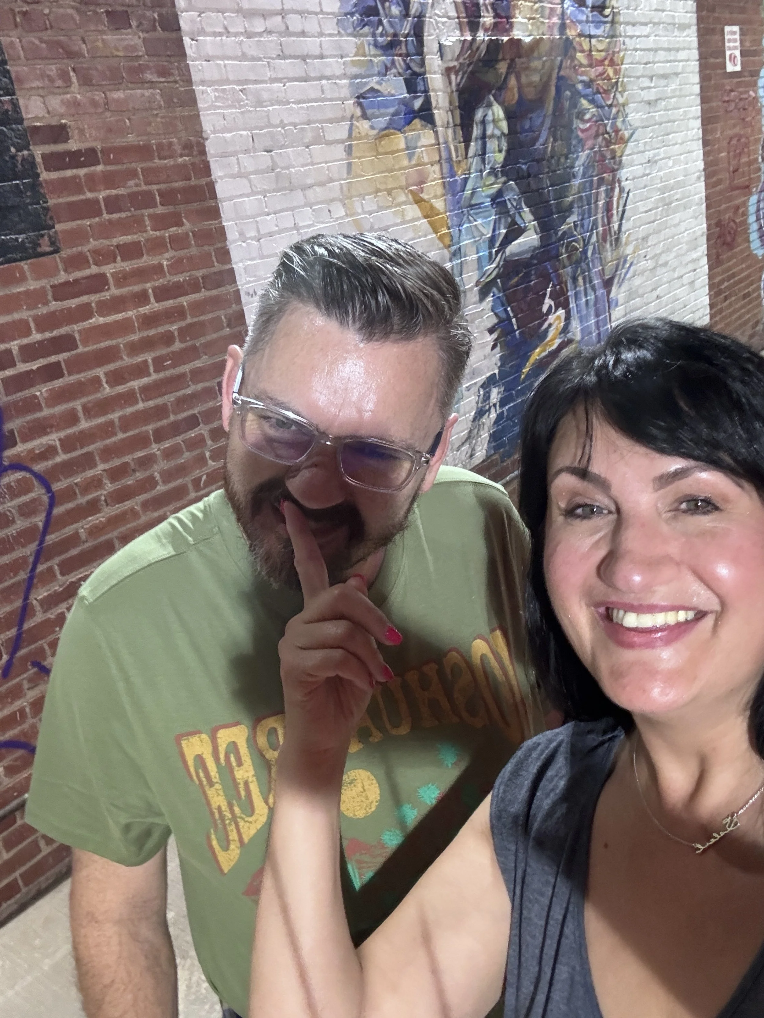 A man with glasses and a woman with black hair smiling, posing together in front of a colorful mural on a brick wall.