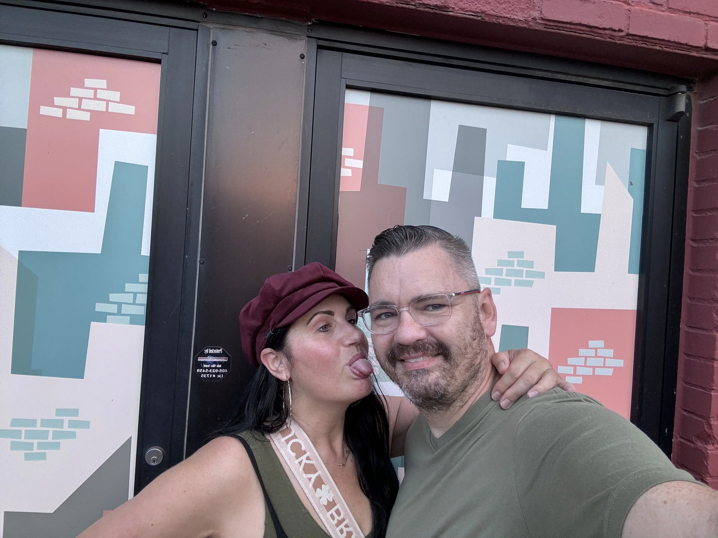 A man and woman taking a selfie outside a building with colorful geometric window decals. The woman is wearing a maroon hat and the man has glasses, both smiling.
