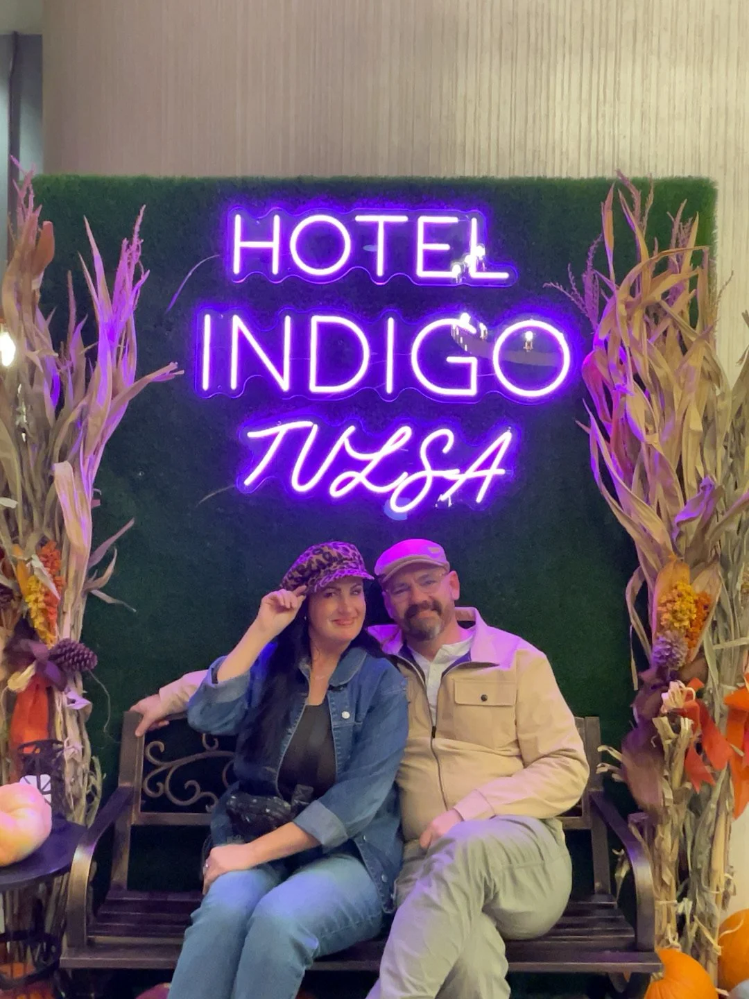 Two people sitting on a bench in front of a green wall with a neon sign that reads 'Hotel Indigo WSSA,' surrounded by fall-themed decorations and dried corn stalks.