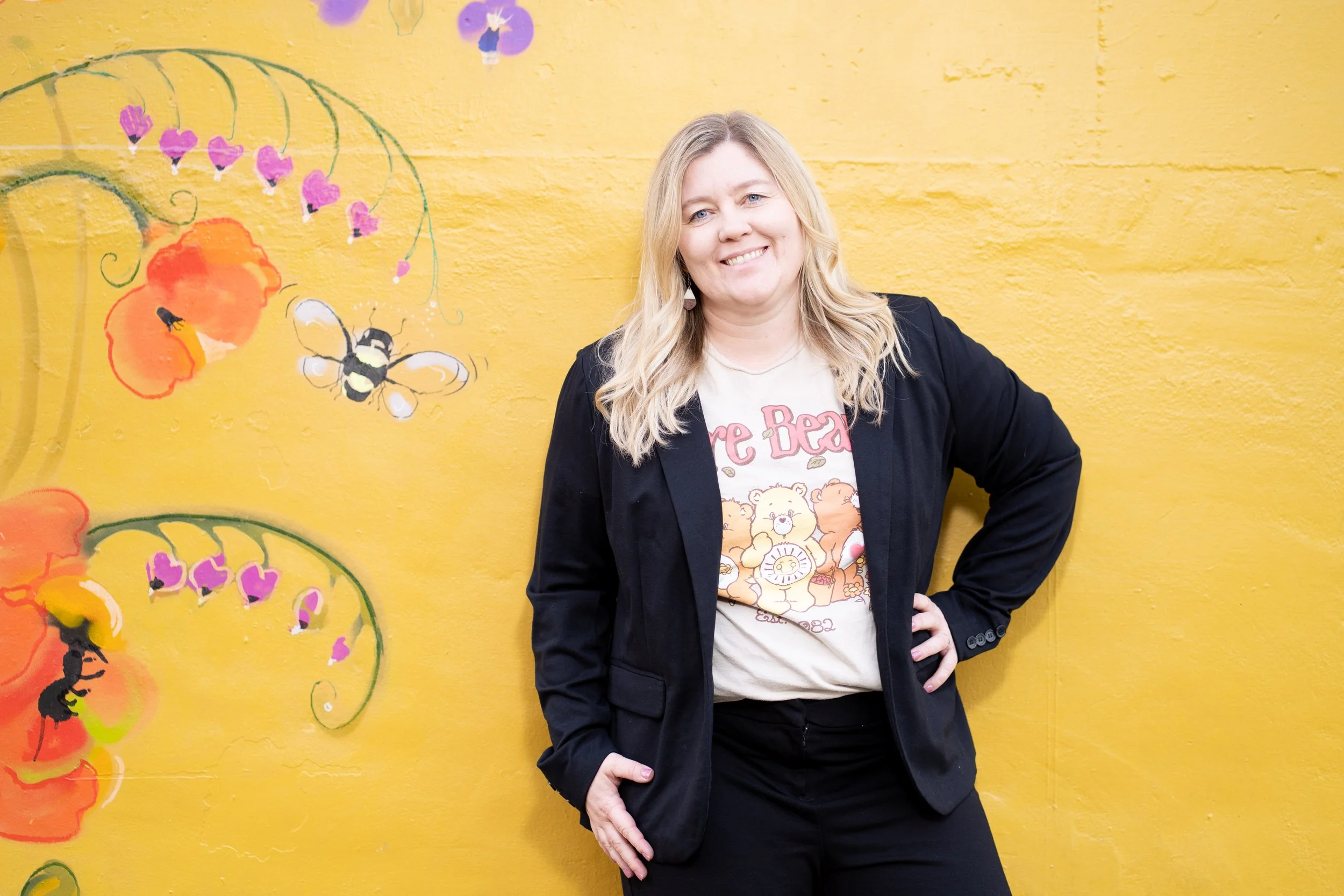 A woman with long blonde hair smiling, wearing a black blazer and a graphic t-shirt featuring cartoon bears, standing in front of a bright yellow wall with colorful, whimsical floral and bee mural artwork.