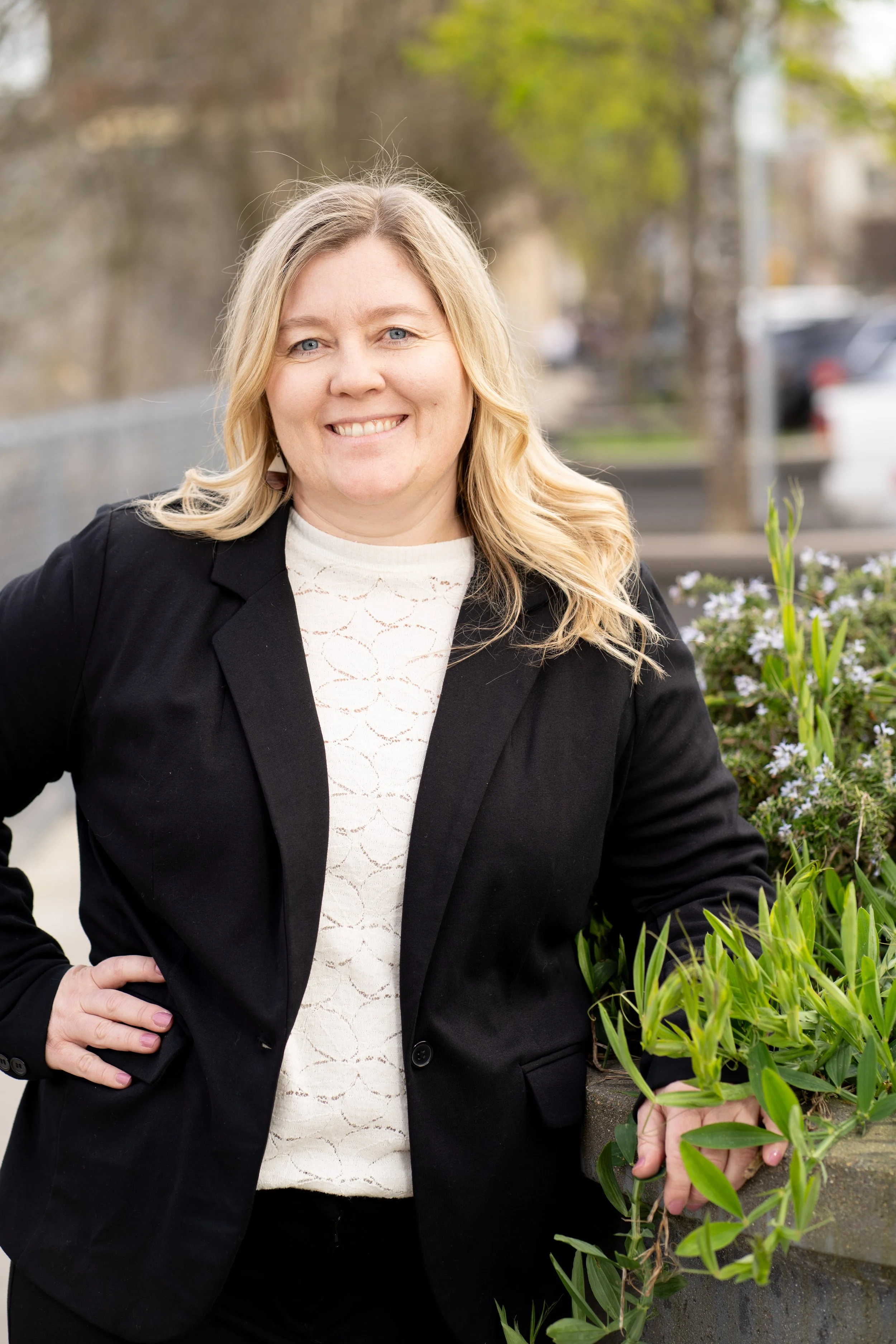 A woman with blonde hair, wearing a black blazer and a light-colored blouse, standing outdoors near a flower bed with green plants and white flowers, with blurred trees and parked cars in the background.