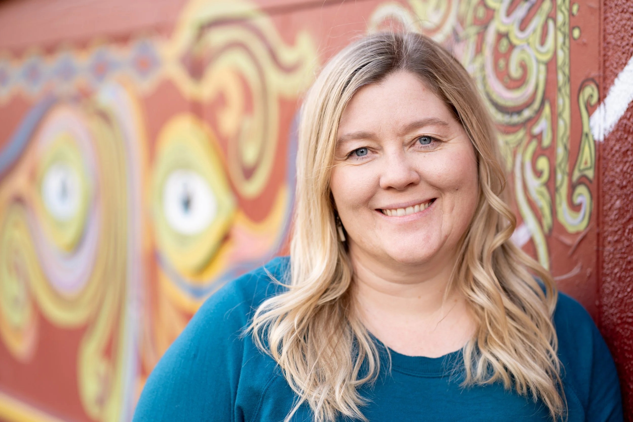 Close-up of a woman with blonde hair and blue eyes smiling in front of a colorful mural.