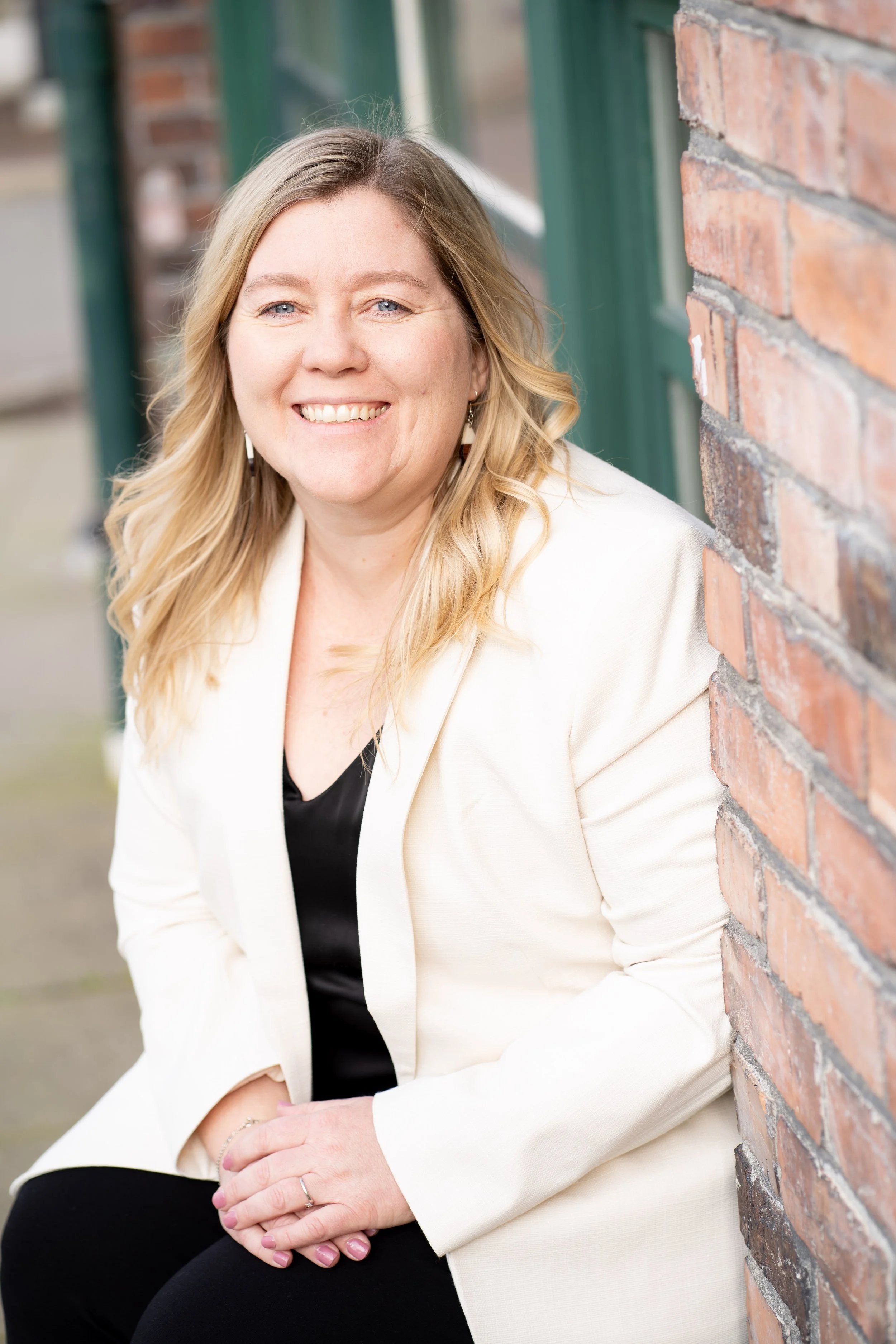 A woman with blonde hair sitting outdoors next to a brick wall, wearing a white blazer and smiling at the camera.