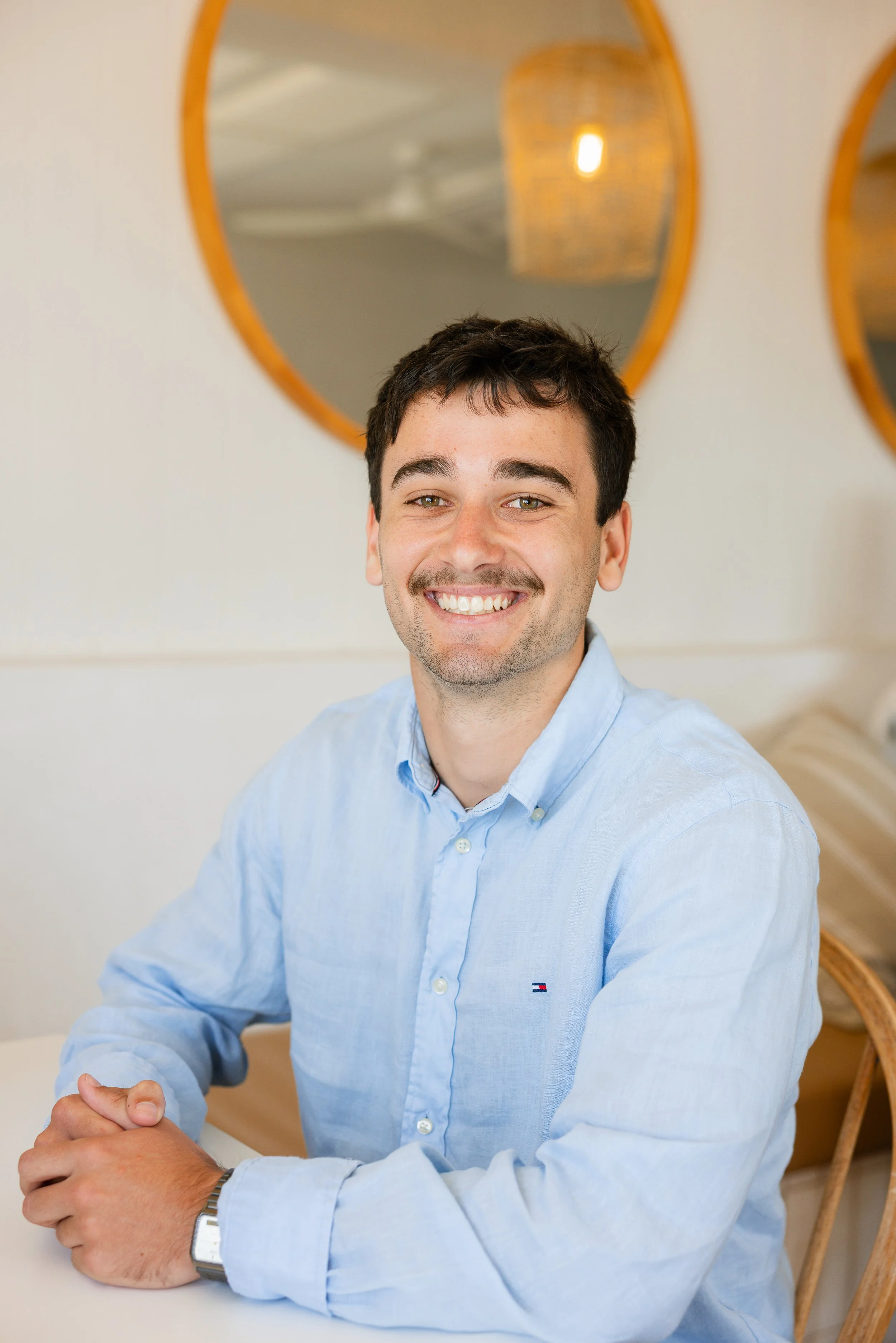 A smiling man with dark hair and a light blue button-up shirt sitting at a table in a room with round mirrors and warm lighting.