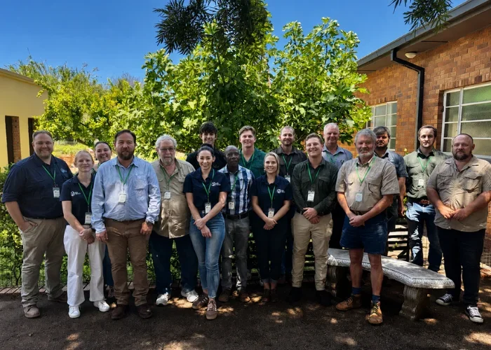Group of 15 people standing outdoors in front of greenery and brick building, smiling for the camera.