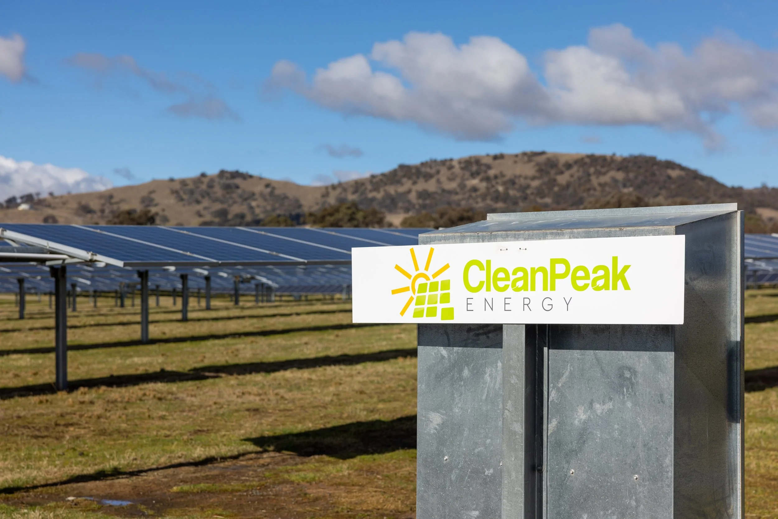 A solar panel farm with a sign that reads 'CleanPeak Energy' in front of the panels, with mountains and a partly cloudy sky in the background.