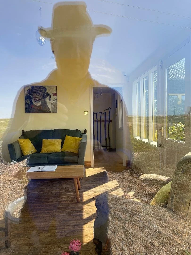 Reflection of a person taking a photo of a living room in a large window with a view of an open field outside. The room has a green sofa with yellow pillows, a wooden coffee table, a piece of artwork on the wall, and a doorway with crutches leaning against it.
