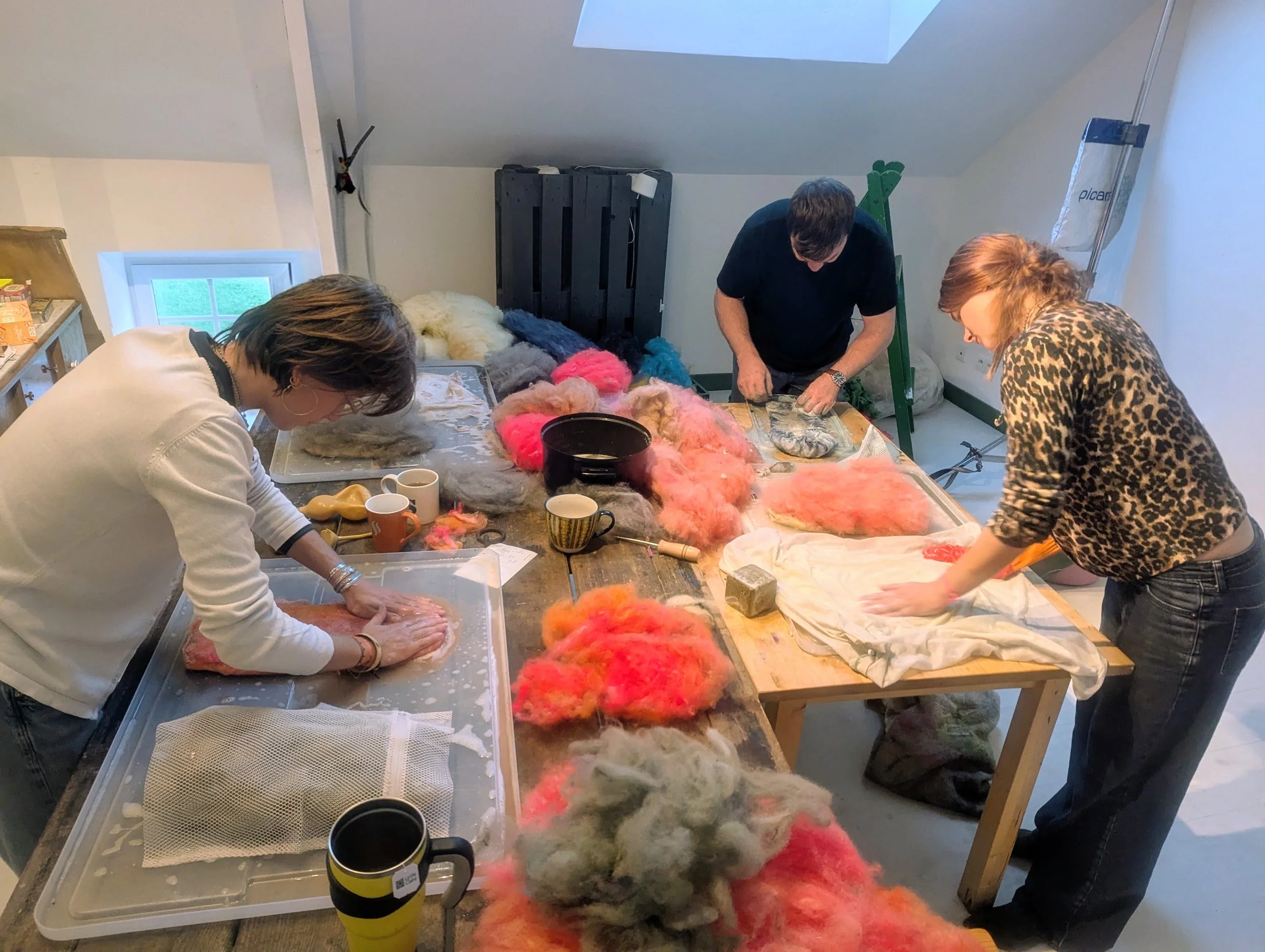 Trois personnes travaillent sur un projet de couture ou de confection de textile dans une pièce éclairée par une fenêtre. La table est encombrée de laine ou de fibres de différentes couleurs. La personne à gauche travaille avec ses mains sur un tissu ou une pièce de laine, la personne au centre manipule quelque chose sur la table, et la personne à droite plie ou prépare un tissu. L'ambiance suggère un atelier ou une activité créative.