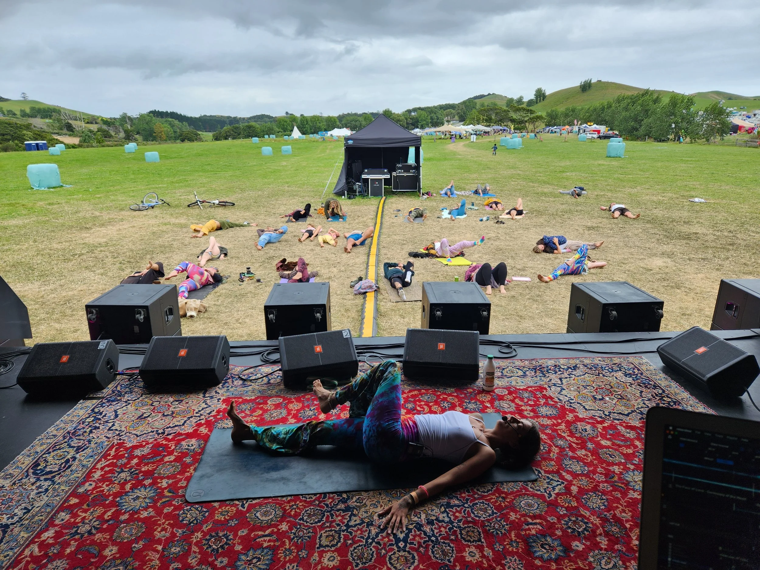 A woman lying on a yoga mat on stage during outdoor yoga class, with participants practicing poses on the grass in front of her, set in a green open field with hills in the background.
