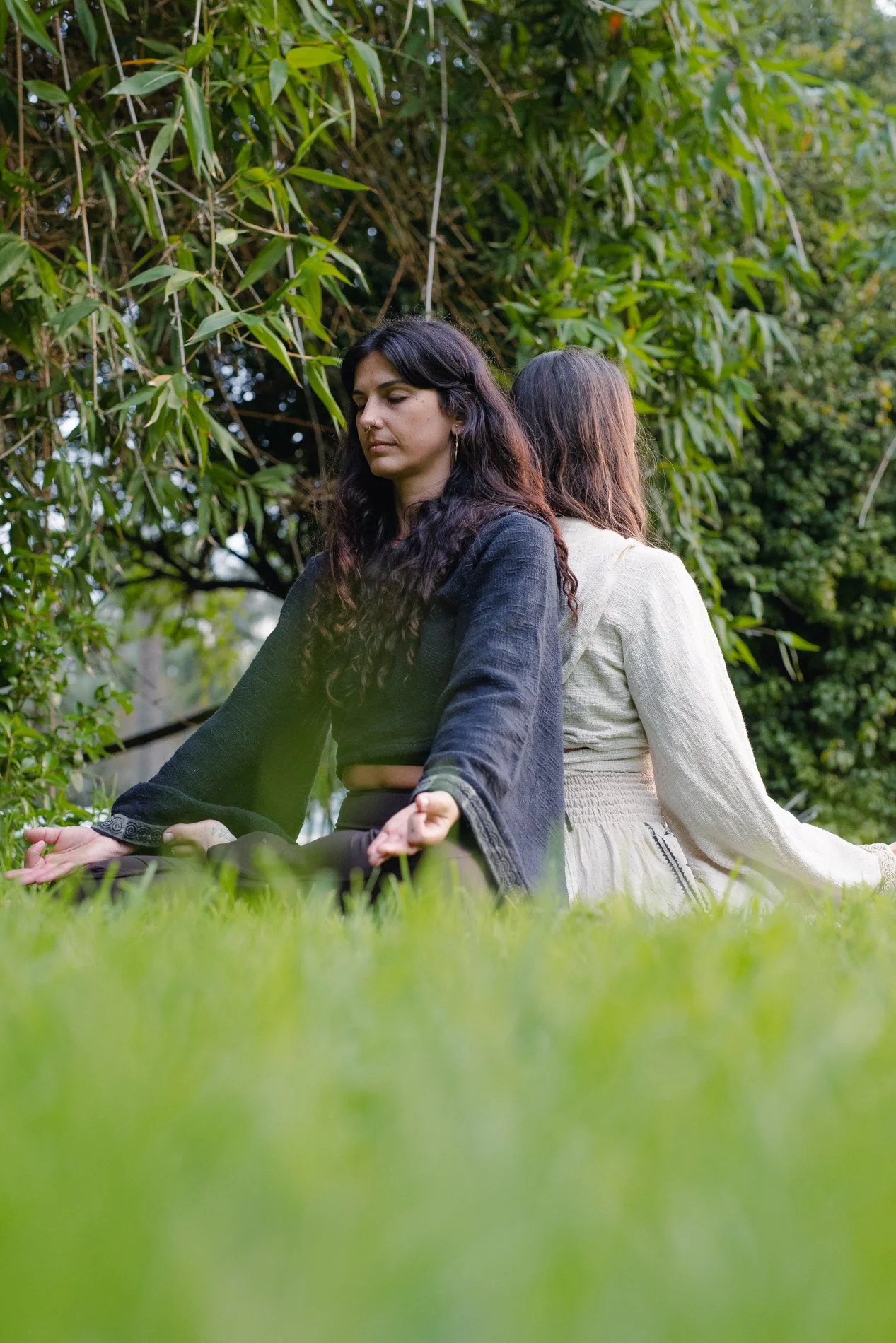Two women sitting on the grass in a meditative pose outdoors, back-to-back, surrounded by lush green foliage.