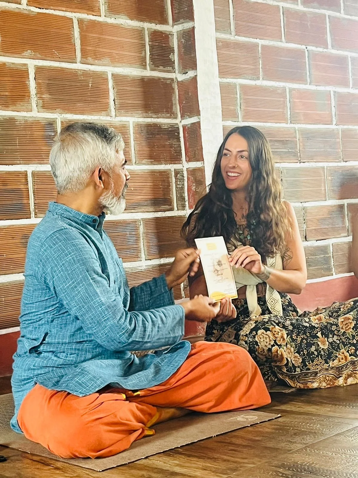 A man and a woman sitting on the floor, engaged in a conversation. The man has gray hair, a beard, and is wearing a blue shirt with orange pants. The woman has dark hair, is smiling, and is holding a booklet or flyer. They are near a brick wall.