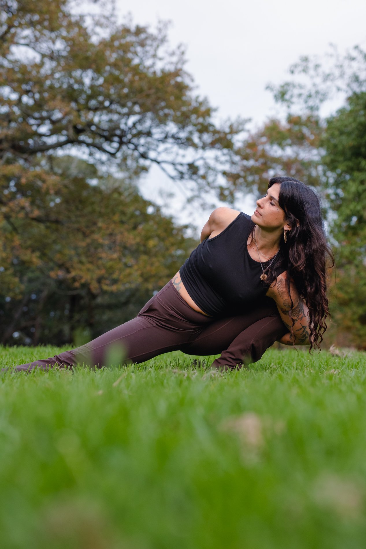 Woman in black tank top and brown pants performing yoga pose on grass in a park with trees in the background.