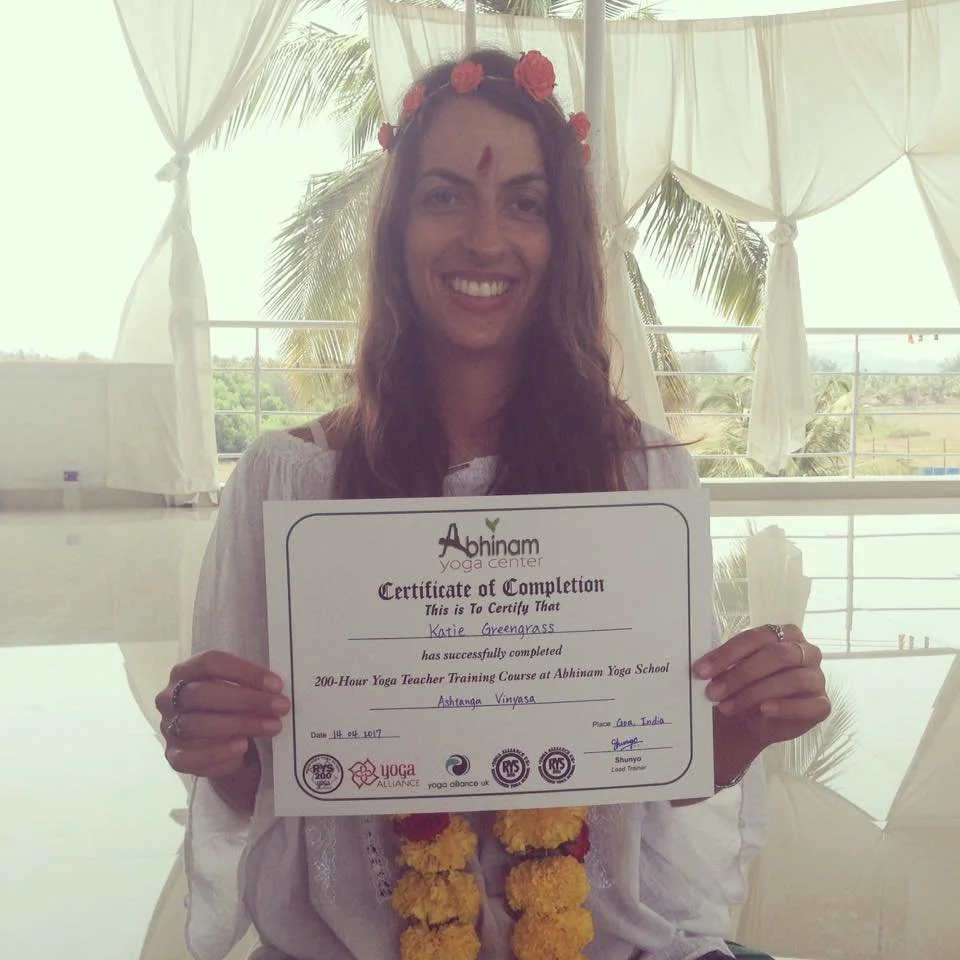 Woman wearing a flower crown holding a certificate of completion from Abhinam Yoga Center, smiling indoors with a background of windows and palm trees.