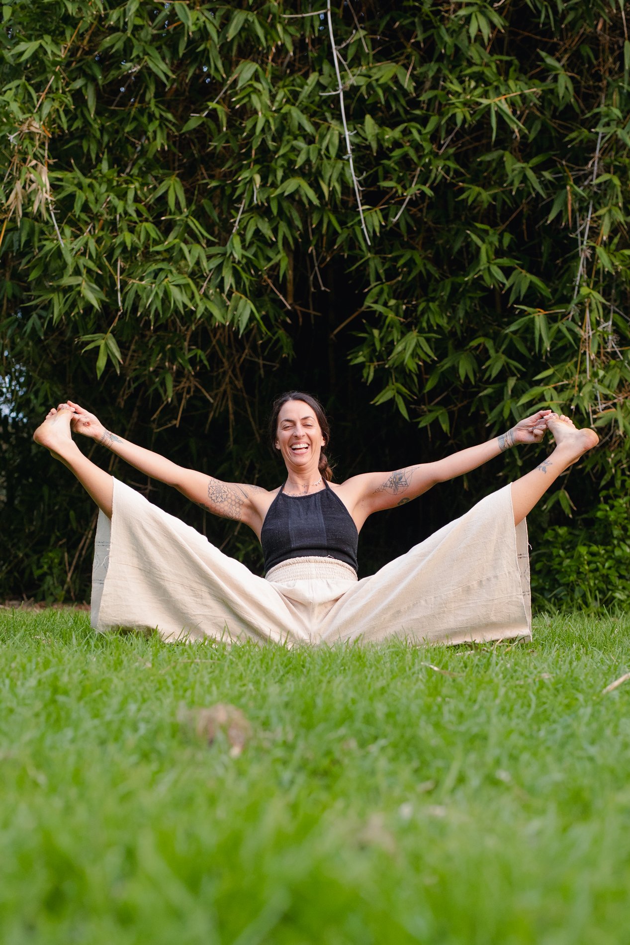 A woman sitting on green grass with her legs spread wide and holding her feet, smiling broadly, in front of tall green leafy bushes.