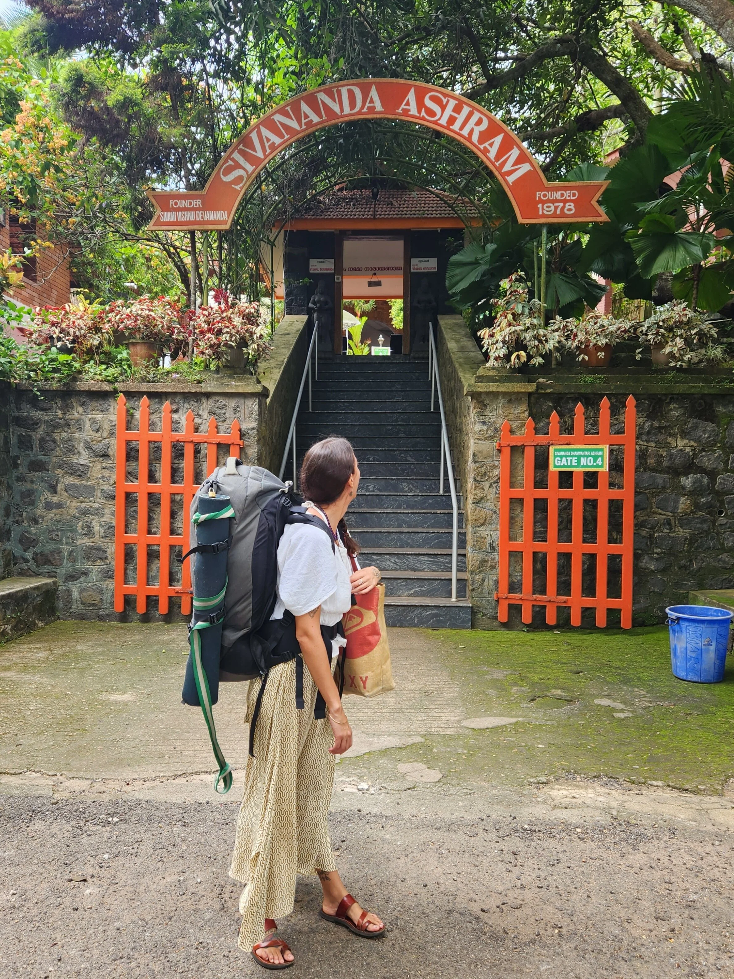 A woman with a large backpack and holding a shopping bag is standing outside the entry gate to Shivanananda Ashram, which has stone steps and orange gates. The sign above the gate shows the ashram was founded in 1978 and is marked as Gate No. 4, surrounded by lush green trees and plants.