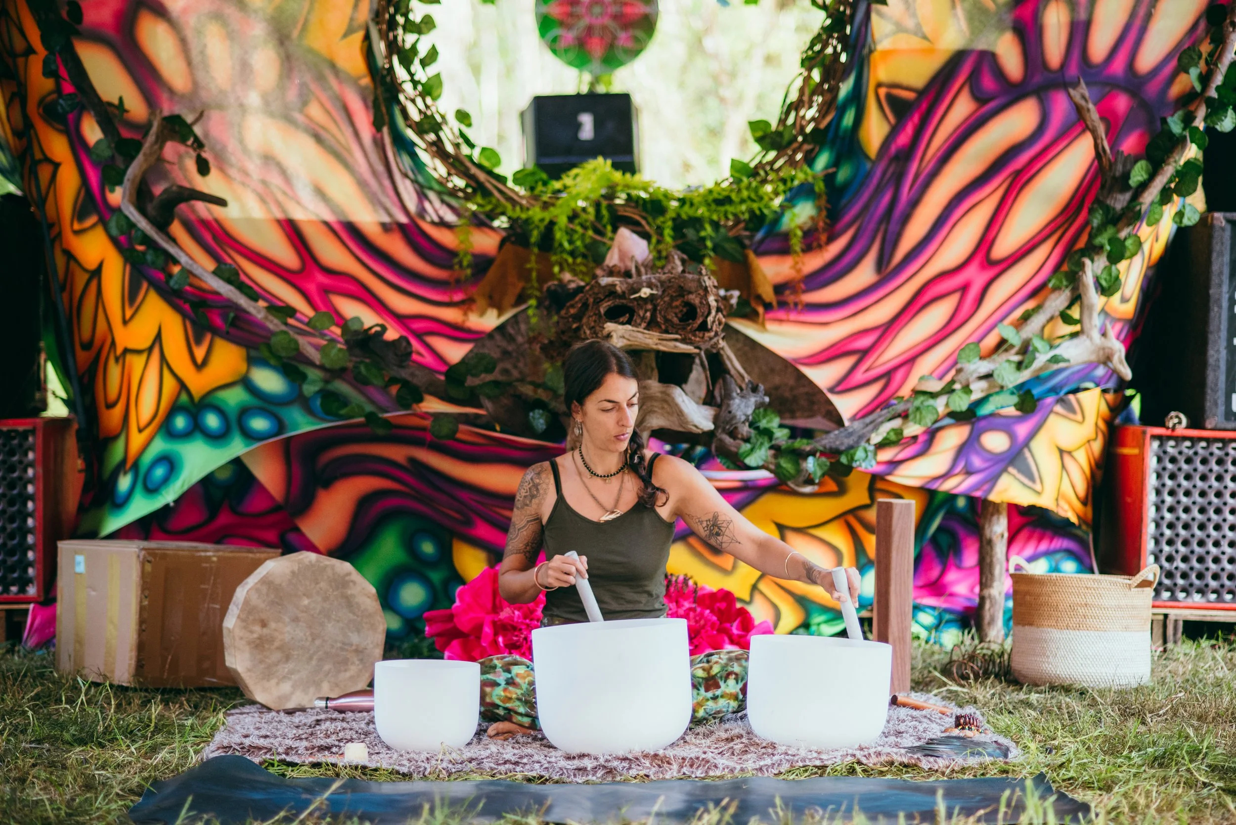 A woman sitting outdoors on a colorful, patterned blanket, playing crystal singing bowls in front of a vibrant, psychedelic backdrop decorated with a dragon skull, driftwood, and greenery.