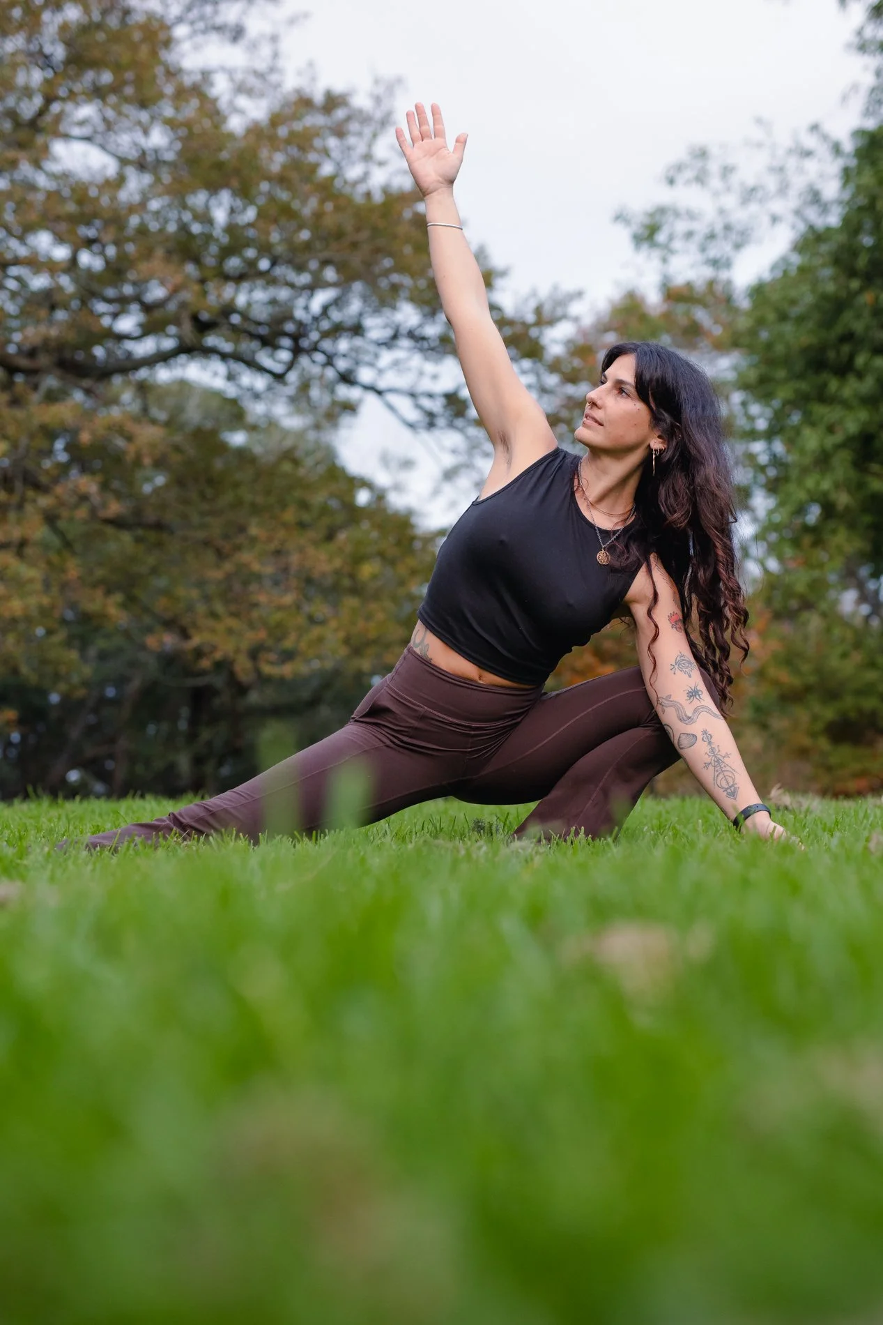 A woman practicing yoga outdoors on grass, doing a side stretch pose with her right arm raised up and her left hand on the ground, surrounded by trees with fall foliage.