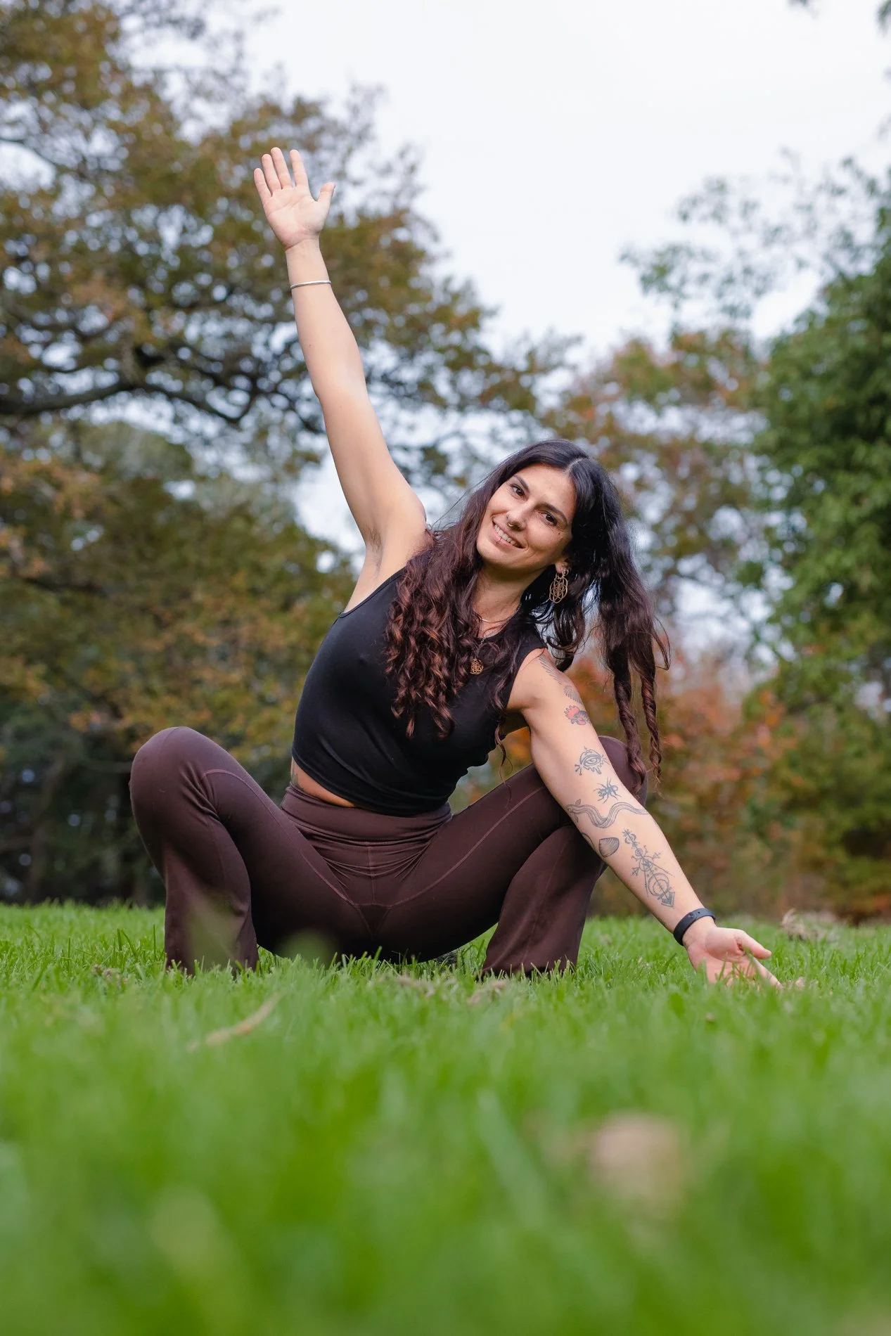A woman practicing yoga outdoors on a grassy field, sitting in a squat pose with one arm raised and smiling at the camera, surrounded by trees with autumn foliage.