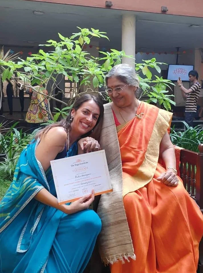 Two women in sarees, one holding a certificate, sitting outdoors near a tree, smiling.