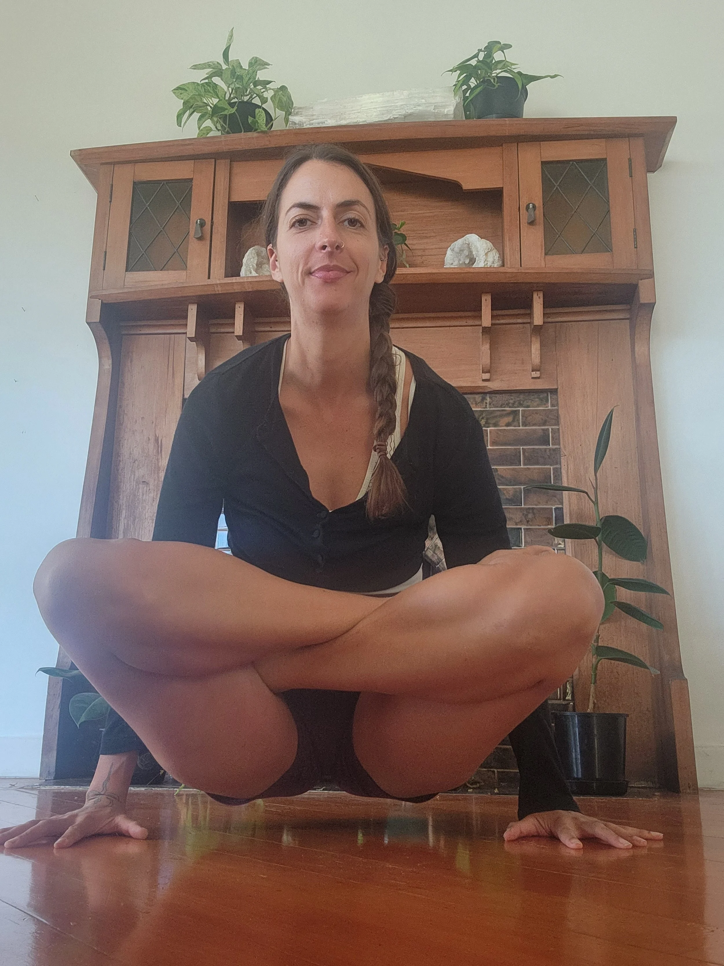 Woman practicing yoga in a low squat position on a wooden floor in front of a wooden fireplace mantel with plants and decorative items.