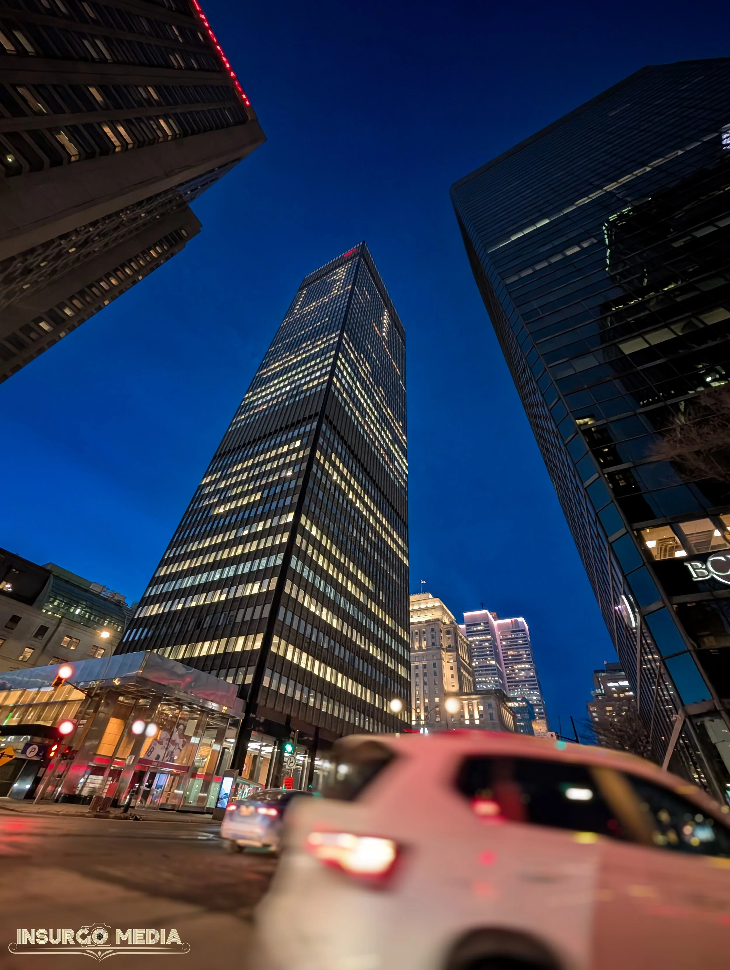 Looking up at tall skyscrapers in a city at dusk, with illuminated windows and a clear dark blue sky.
