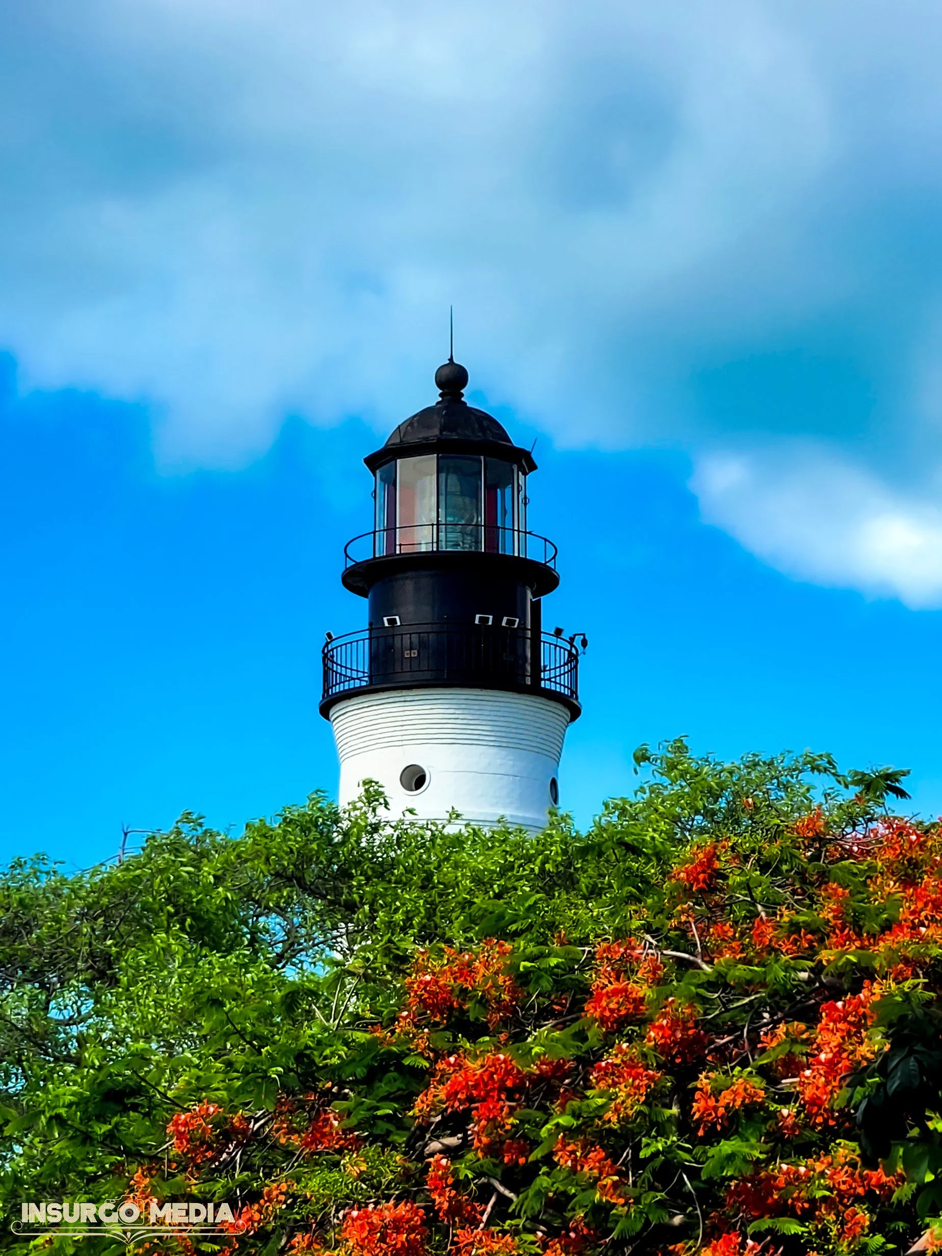 A lighthouse standing tall behind green and orange flowering trees against a partly cloudy blue sky.