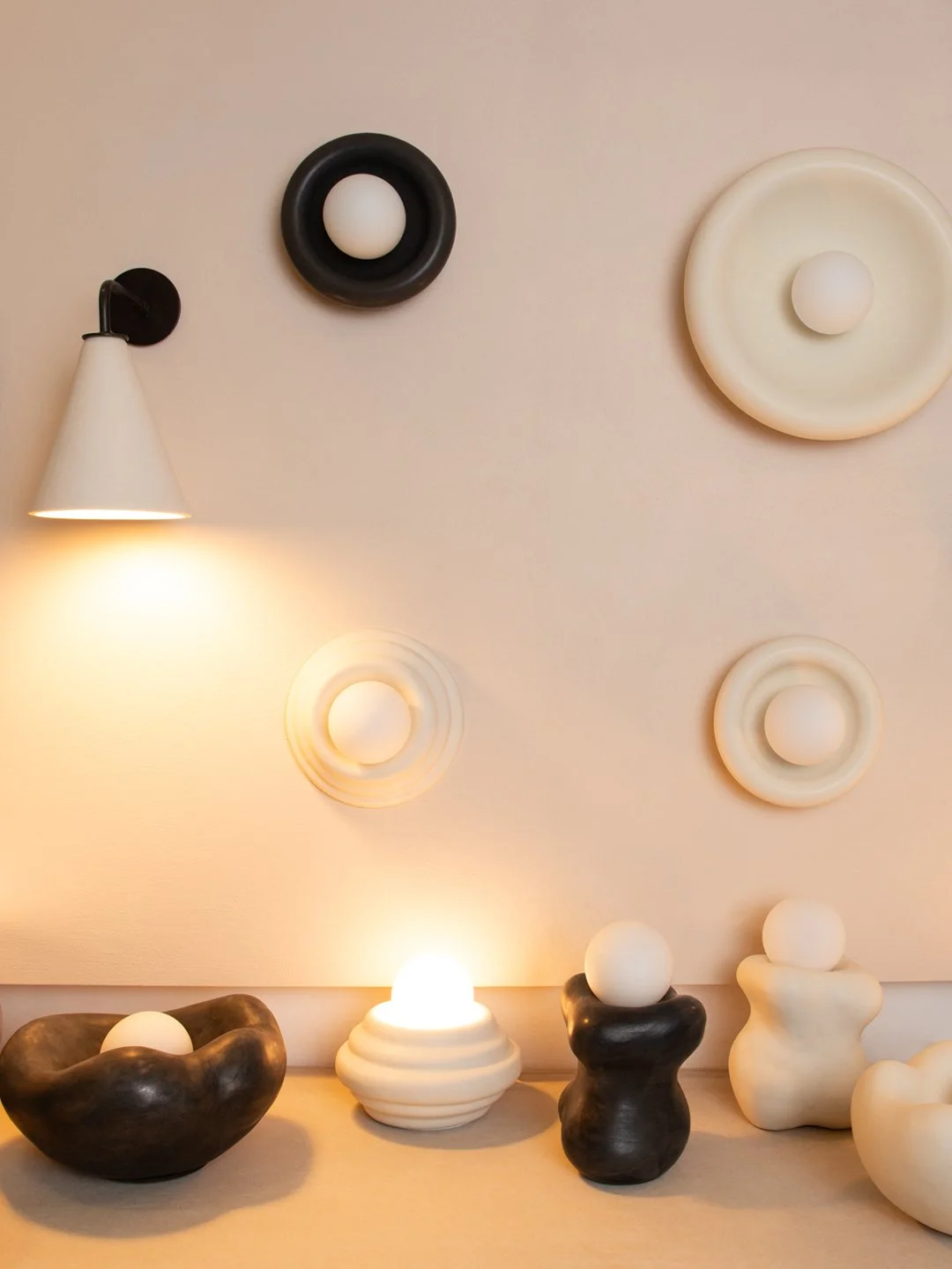 Decorative wall art with multiple circular wall sculptures and a white wall-mounted light, arranged on a beige wall above a surface with abstract black and white ceramic sculptures and a glowing lamp.
