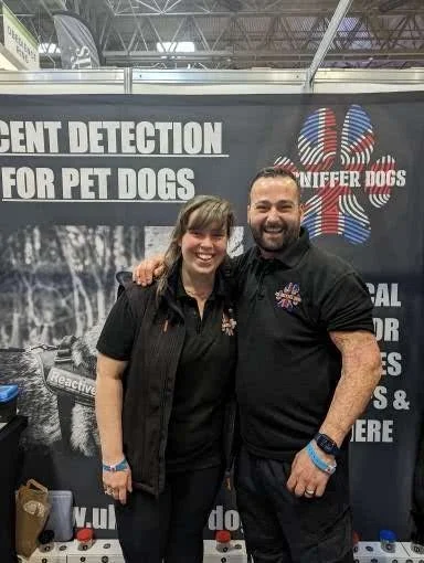Two people, a woman and a man, standing together at a booth promoting pet dog scent detection. They are smiling and wearing black shirts with a logo. The booth has a banner with the words 'Scent Detection for Pet Dogs' and an American flag-themed paw print logo.