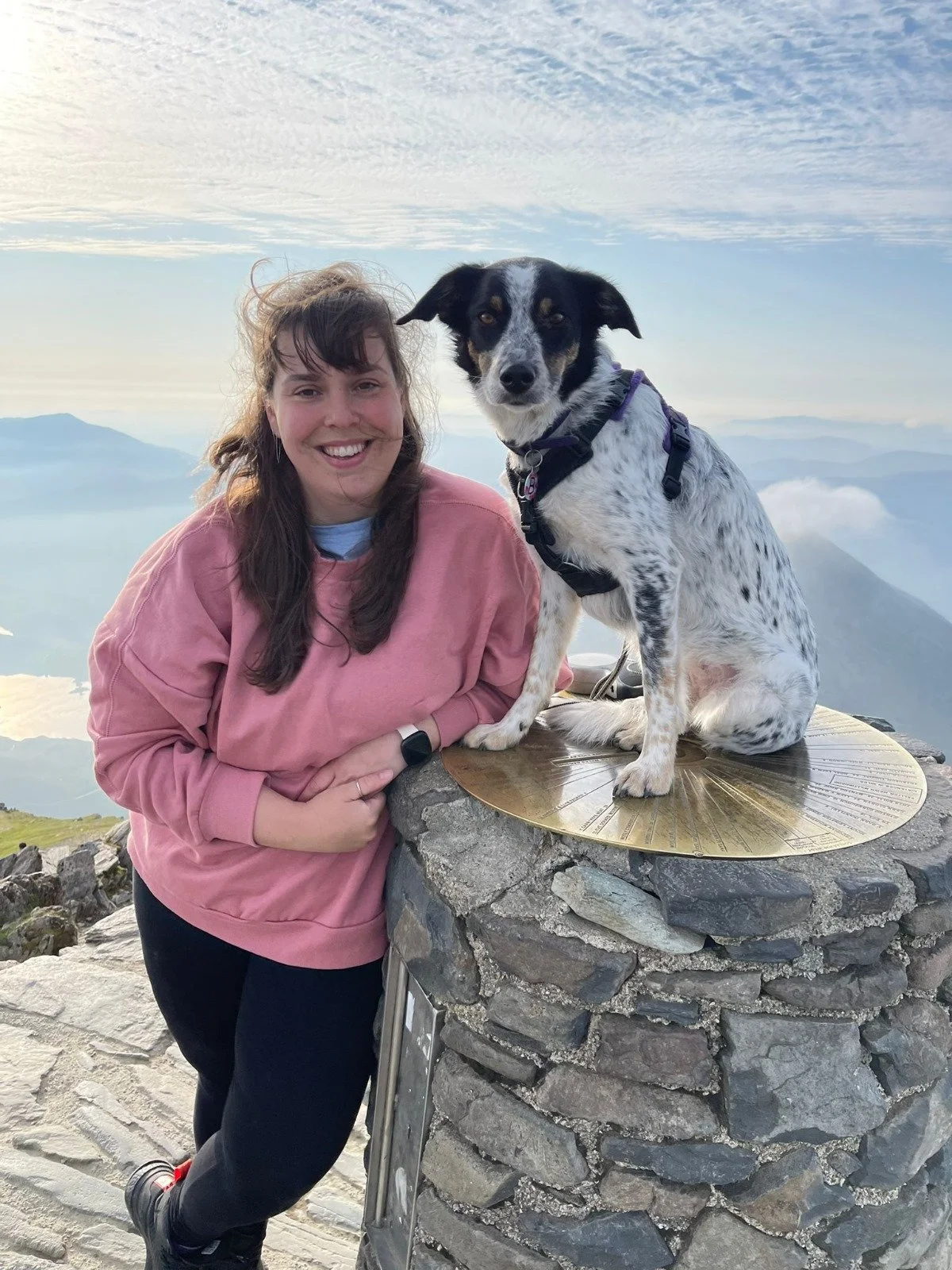 A woman smiling in a pink sweatshirt standing next to a black and white dog sitting on a stone pedestal, with a mountain landscape and sky in the background.