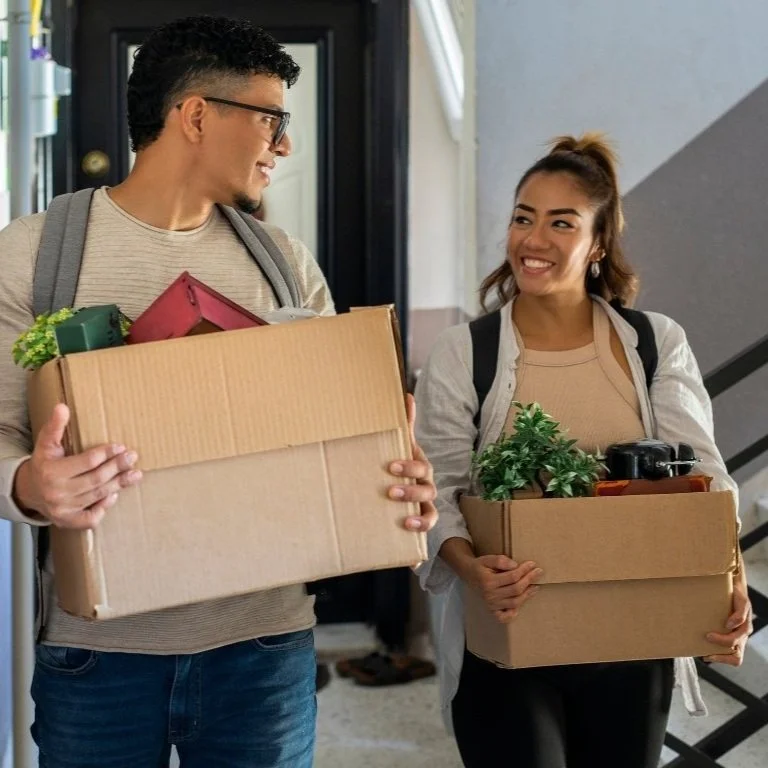 A man and woman carrying cardboard boxes while standing at a staircase, smiling and looking at each other in a building.