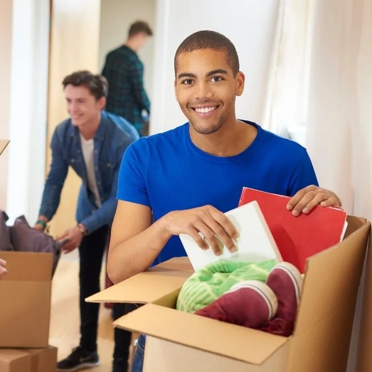 A young man smiling while packing a cardboard box with books and clothes. Two other people are visible in the background, also preparing boxes.