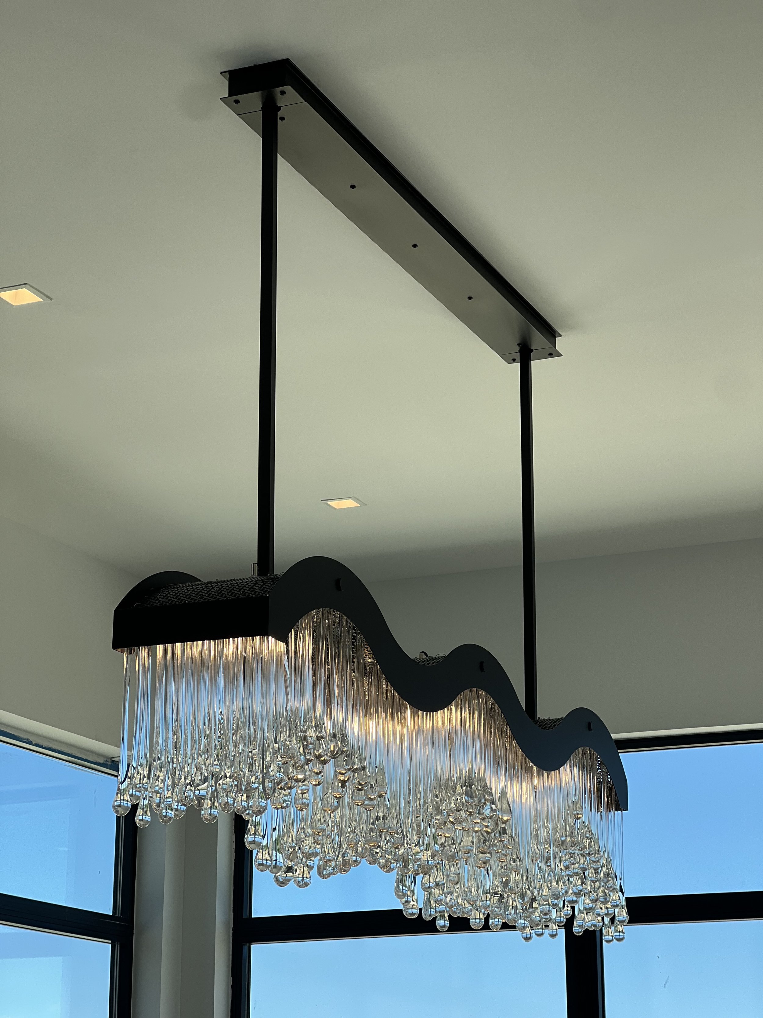 Modern chandelier with a black wavy top and cascading glass drops, hanging from a black rectangular ceiling mount with two supporting rods, in a room with large windows and natural light.