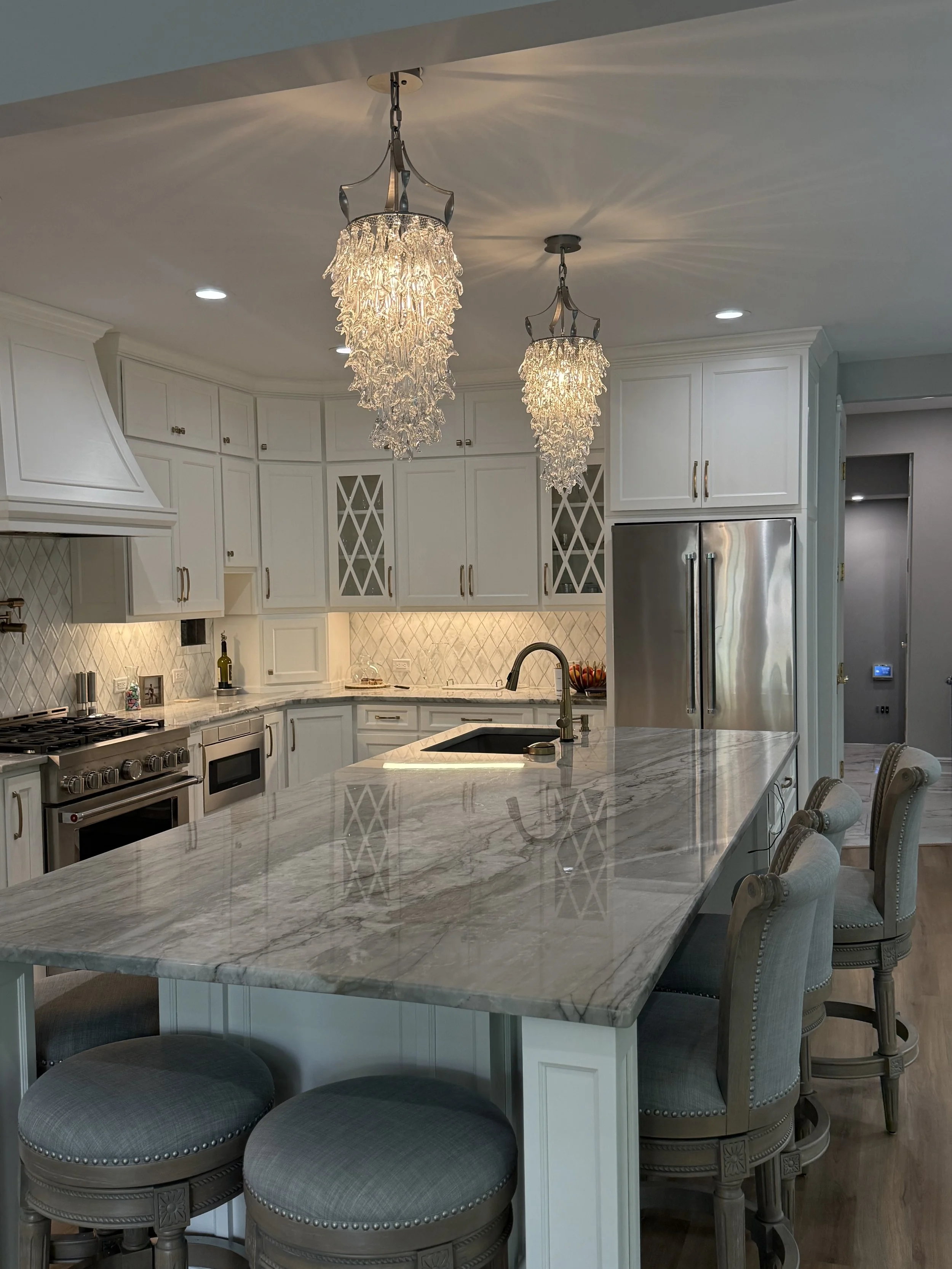 A modern kitchen with a large marble island, white cabinets, stainless steel refrigerator, and elegant chandeliers hanging from the ceiling.