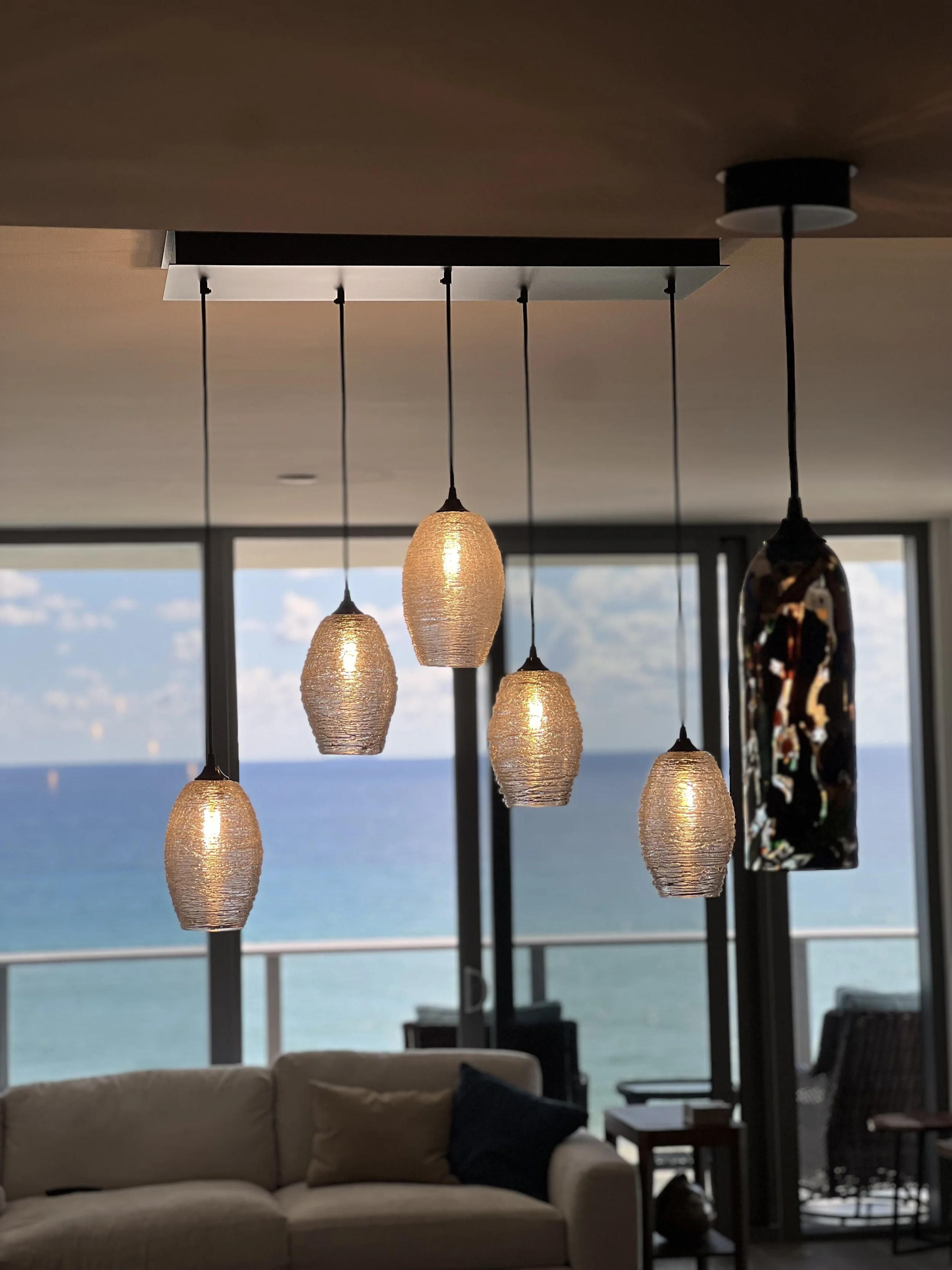 Modern hanging pendant lights in front of a large window with a view of the ocean and blue sky.