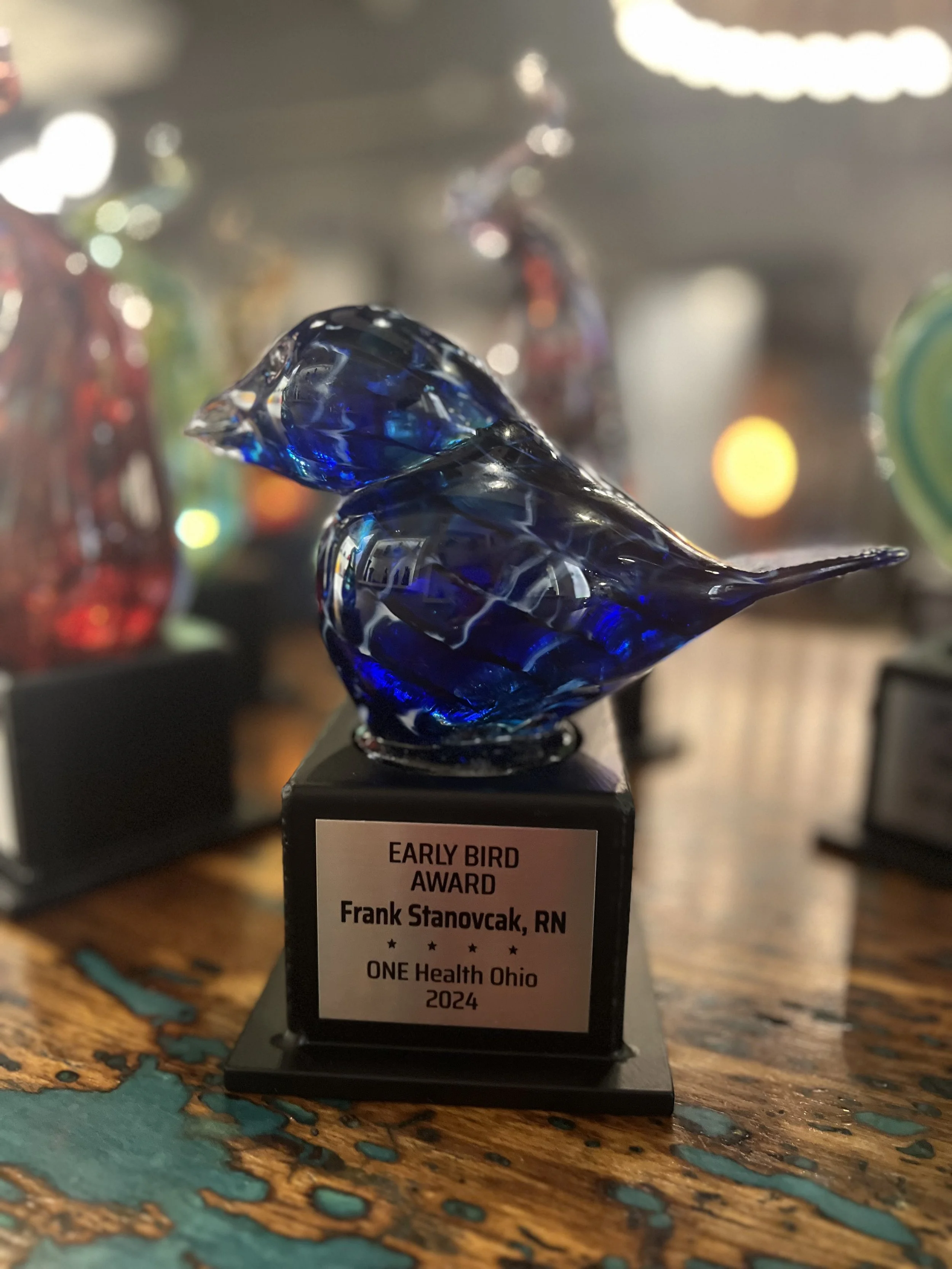 Blue glass bird trophy with a plaque that reads, 'Early Bird Award, Frank Stanovcak, RN, ONE Health Ohio, 2024' on a wooden table.