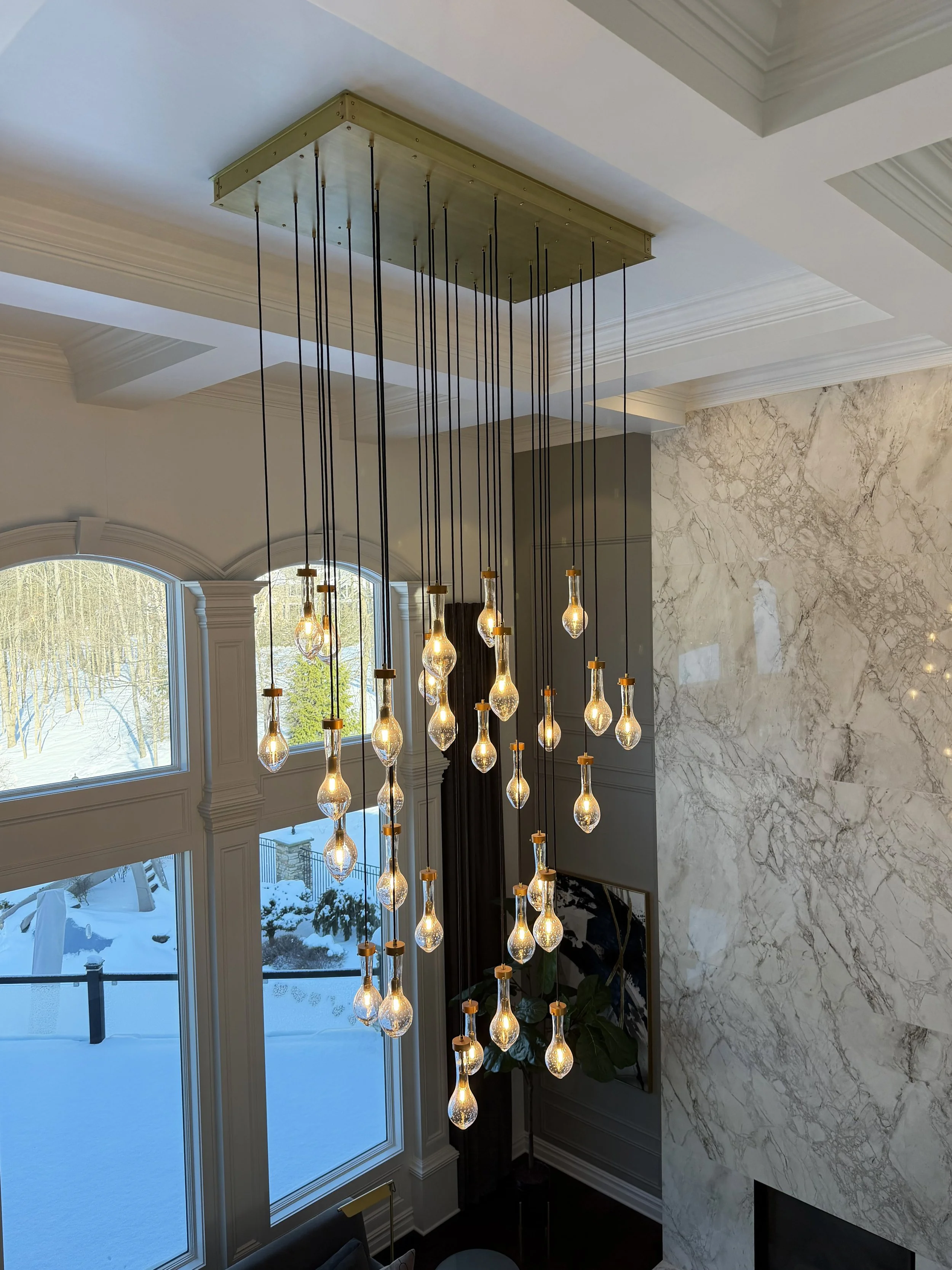 Modern chandelier with multiple illuminated teardrop-shaped glass bulbs hanging from black cords over a living room with large windows, a marble wall, and snowy landscape outside.