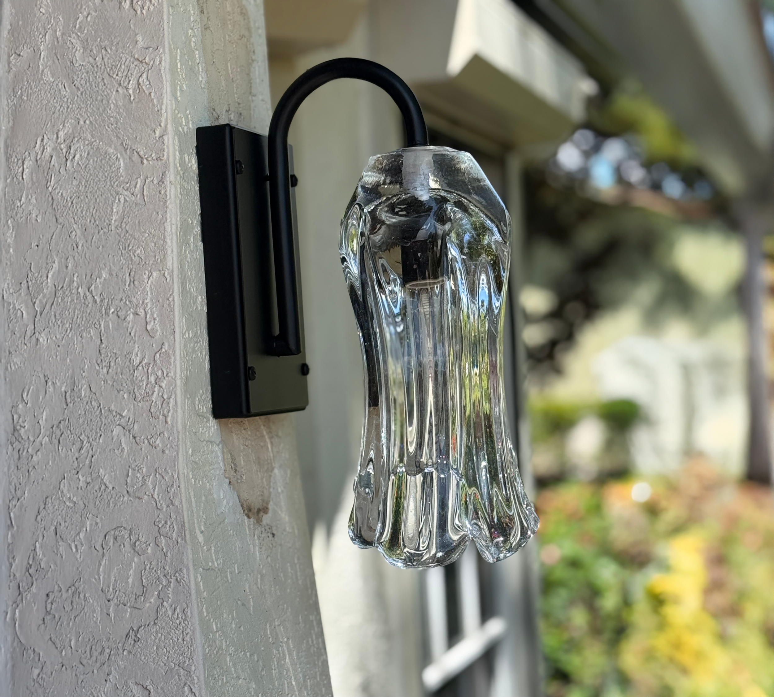 Close-up of a glass pendant outdoor wall light fixture attached to a textured wall with greenery and a blurred tree in the background.