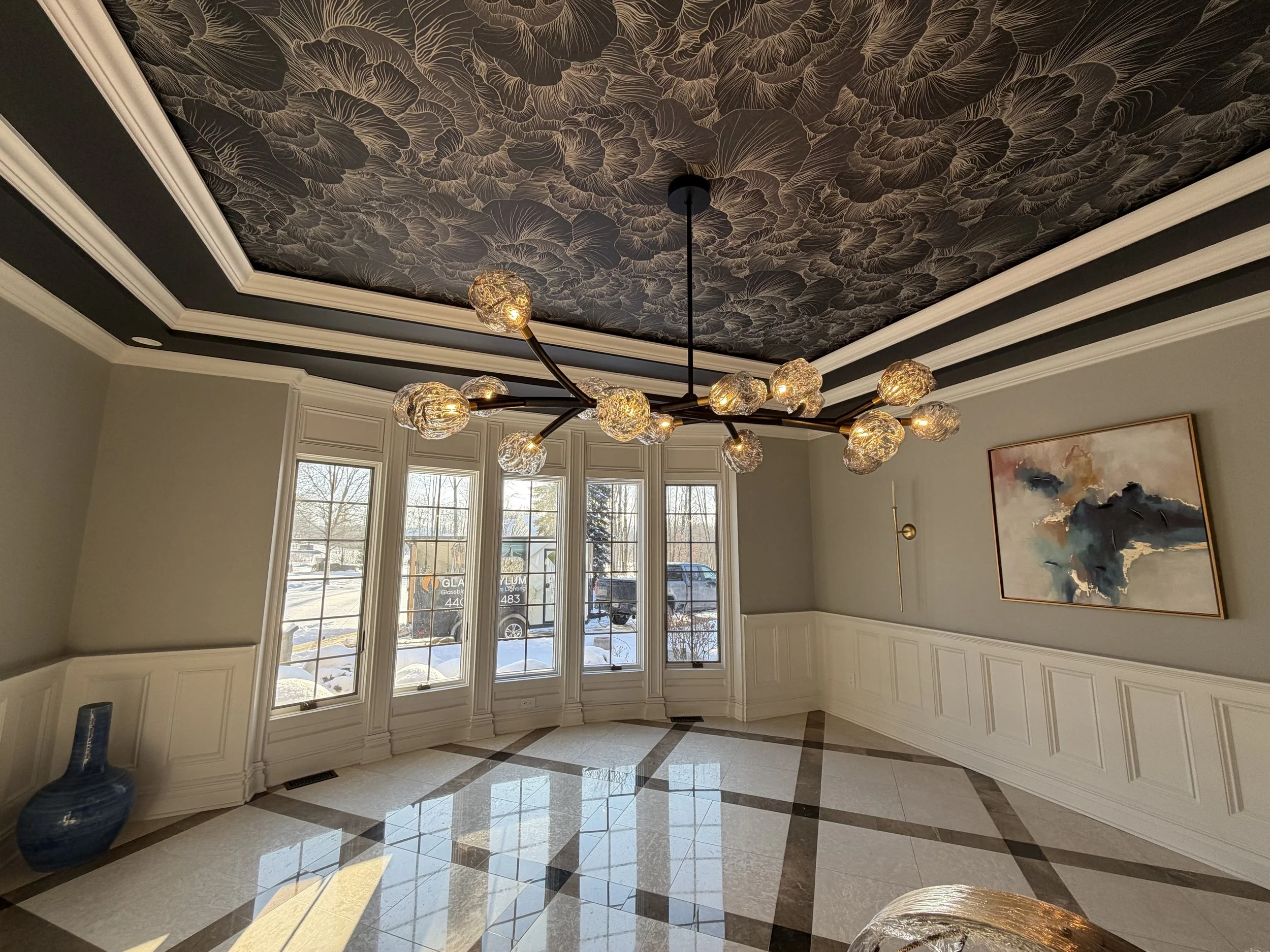 A modern, empty room with large bay windows, patterned tiled floor, wainscoting, a contemporary chandelier with multiple glass globes, and a wall-mounted abstract painting.