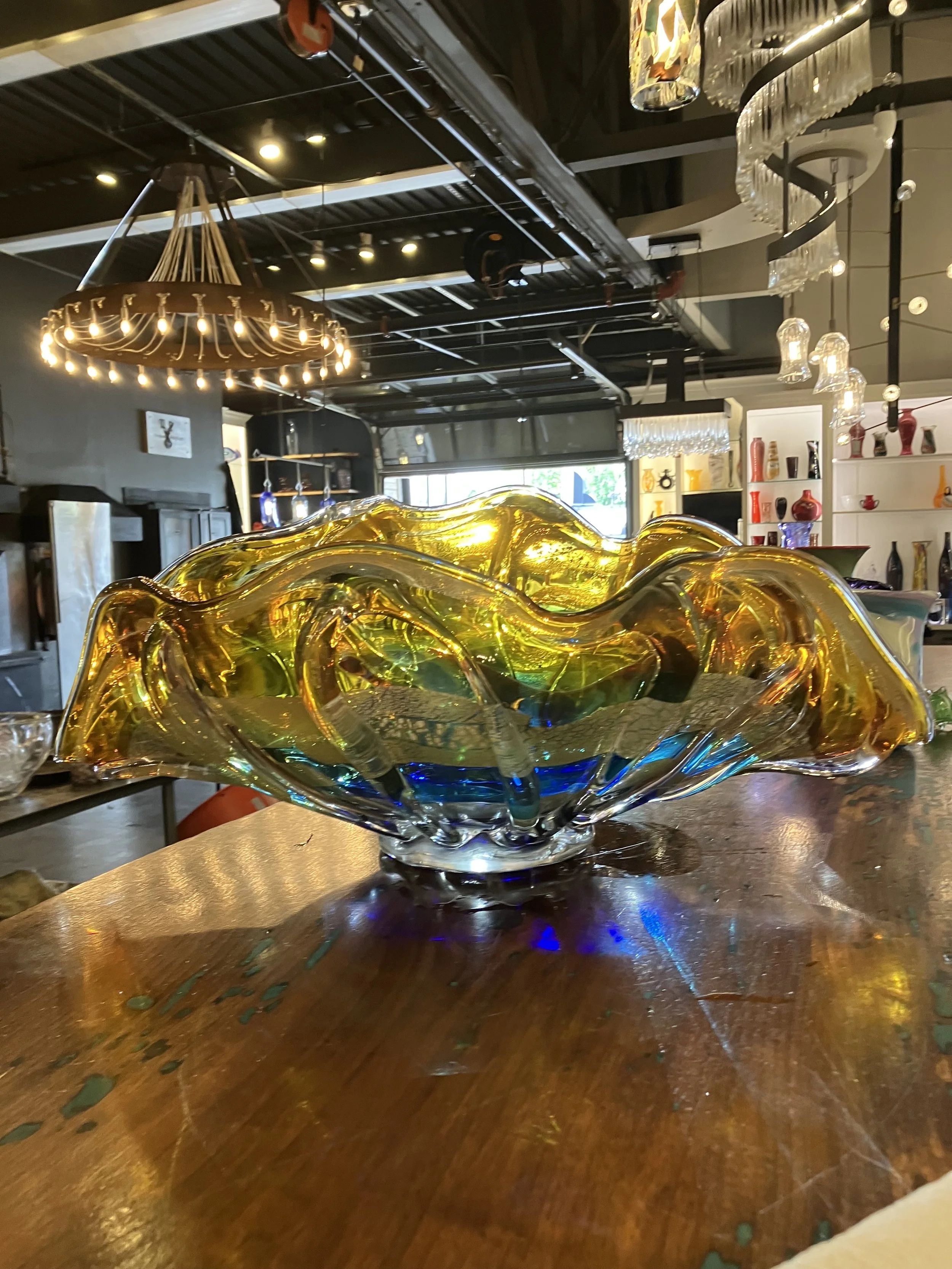 Colorful glass art piece on a wooden table inside a modern studio, with decorative lighting and shelves with vases in the background.