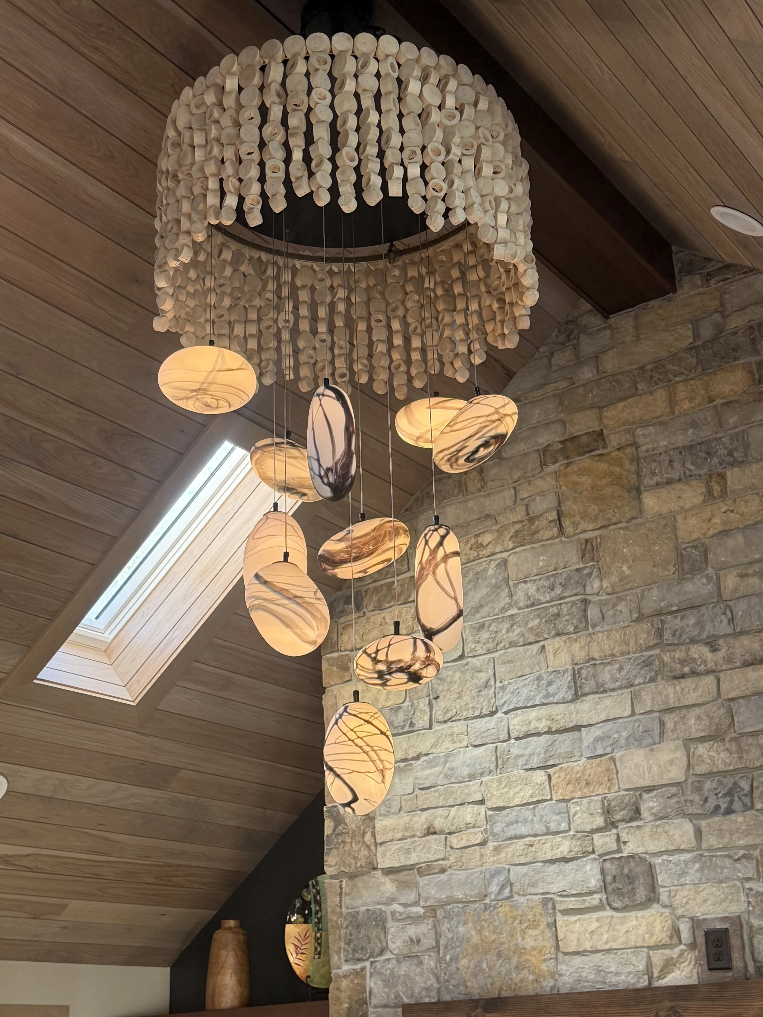A large chandelier with cream-colored wood beads and hanging marble-like lampshades in a modern interior ceiling, next to a stone wall and a skylight window.