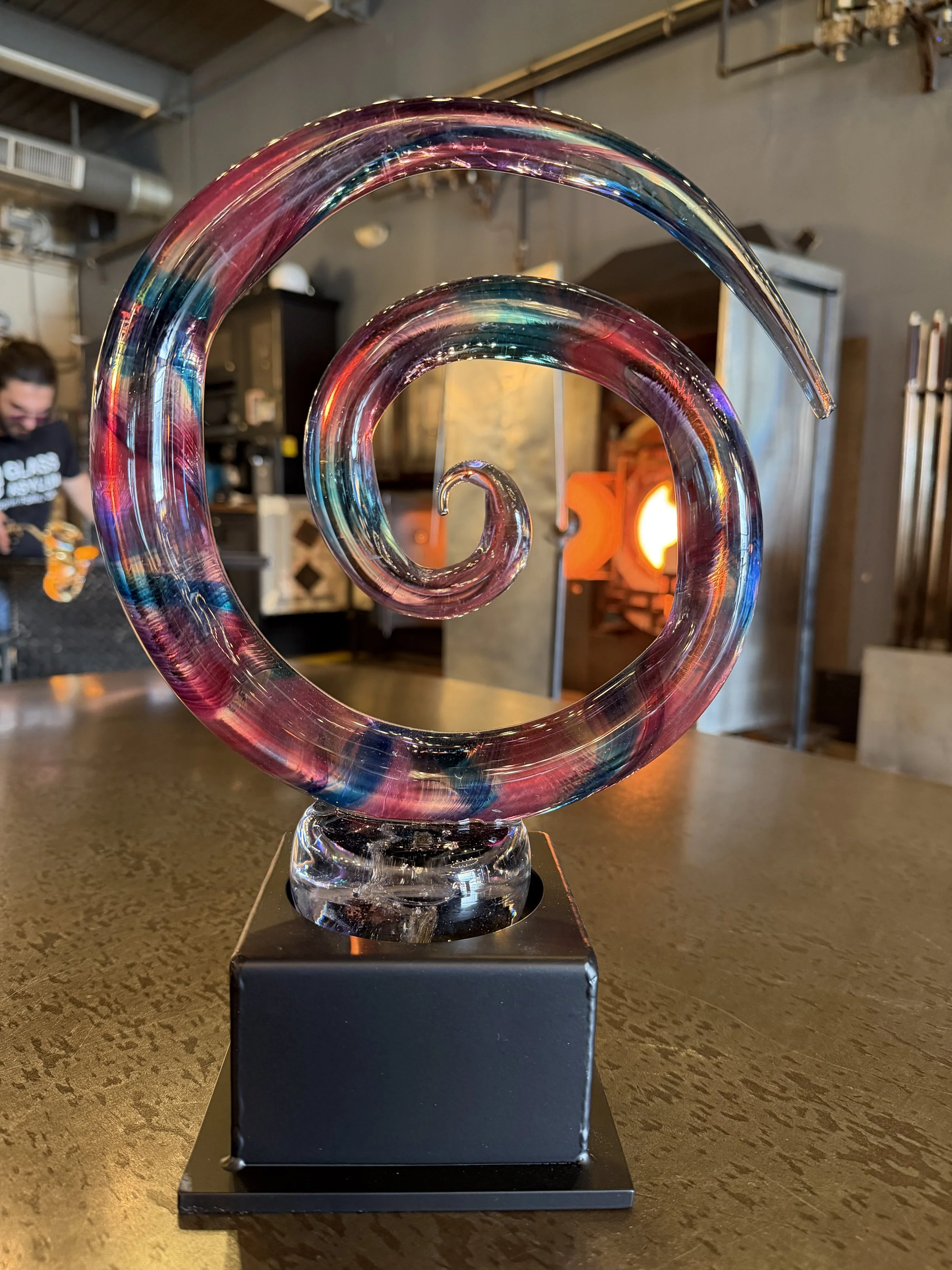 Colorful glass spiral sculpture on black base inside a modern workspace with furnace in background.