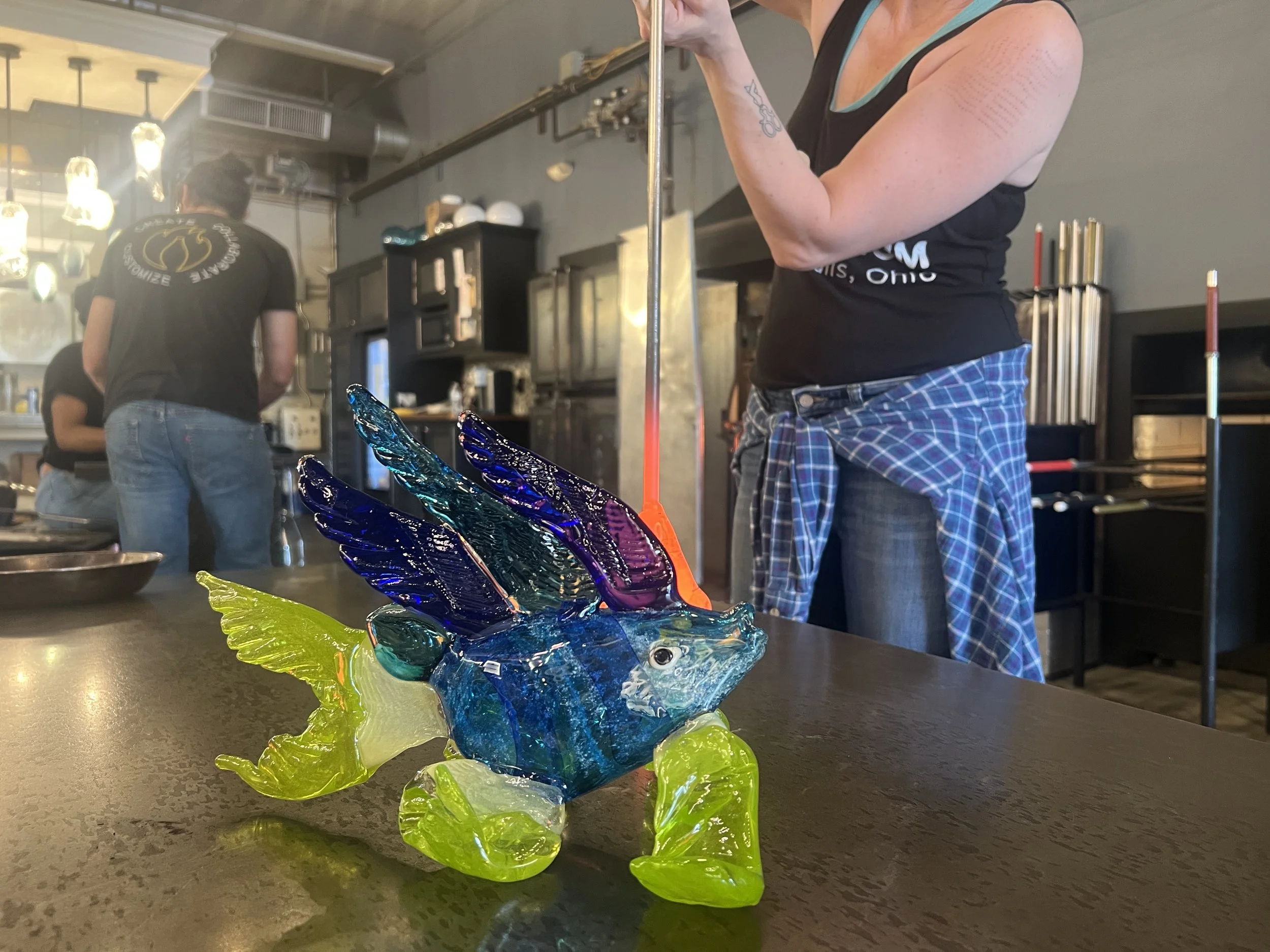 Colorful glass sculpture of a fish with wings placed on a dark table in front of a person blowing glass.
