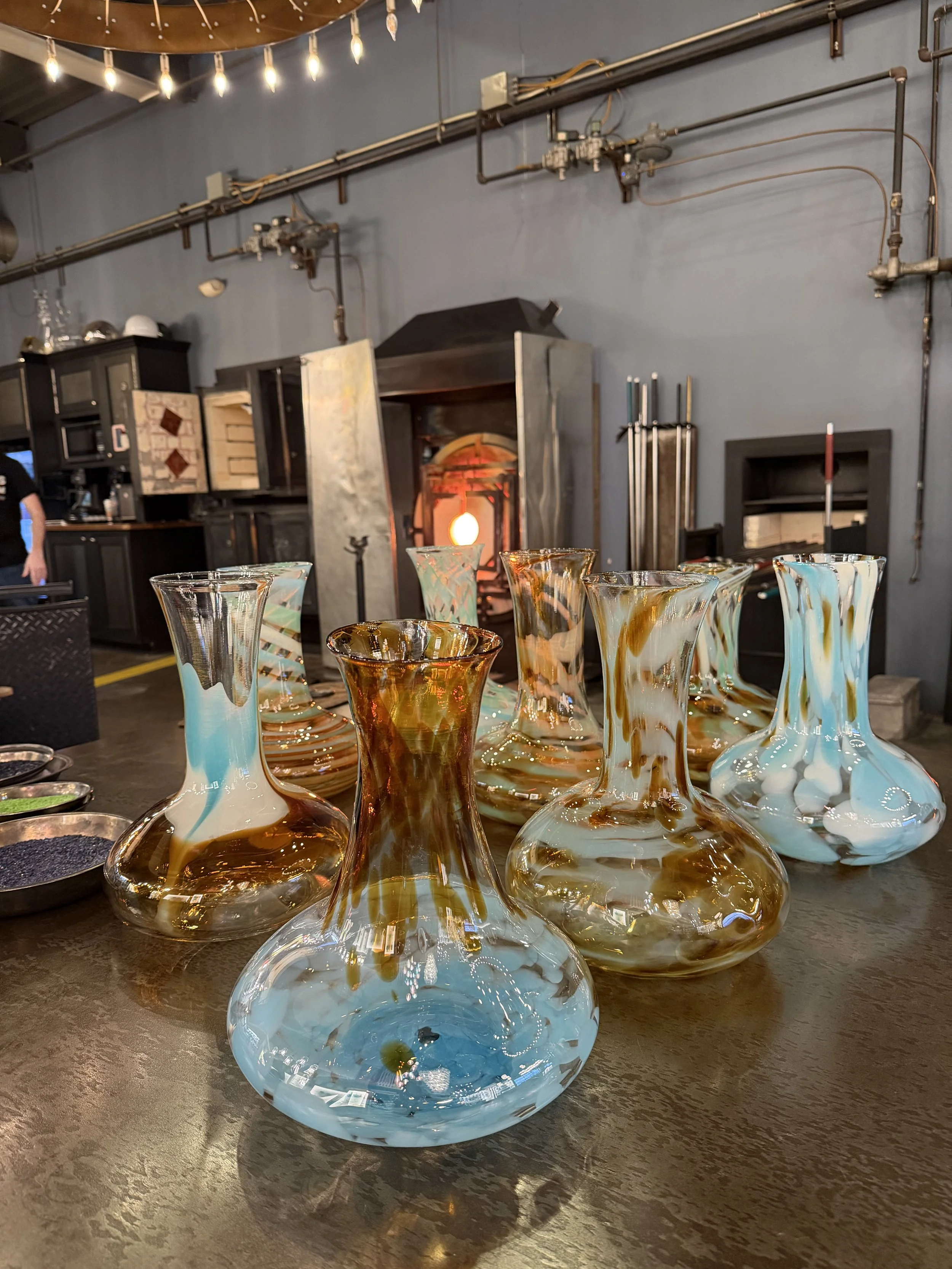 Colorful glass decanters on a table in a glassblowing workshop, with a furnace in the background.