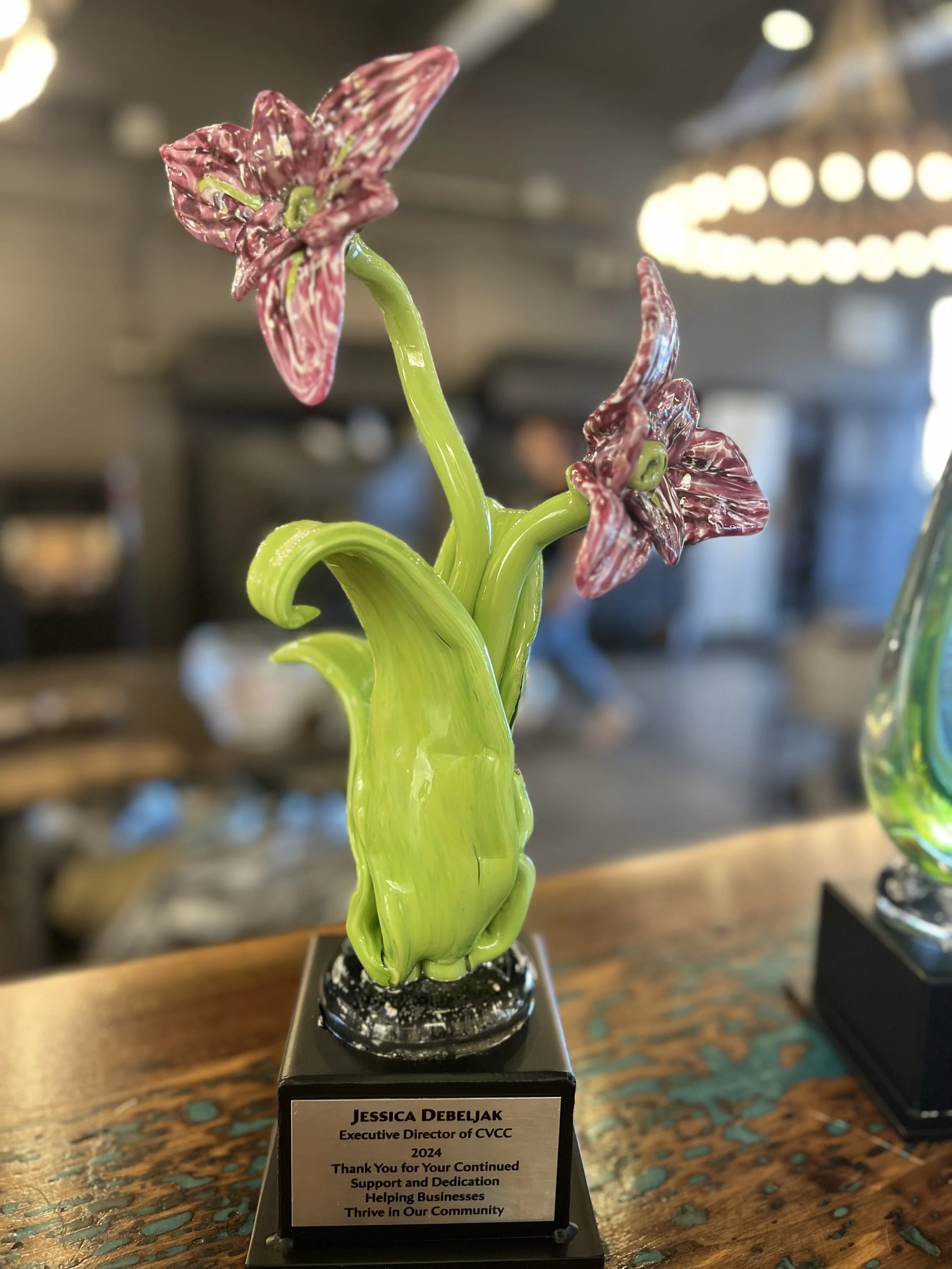 Glass sculpture of a pink and white striped flower with green leaves, displayed on a black base with a plaque.