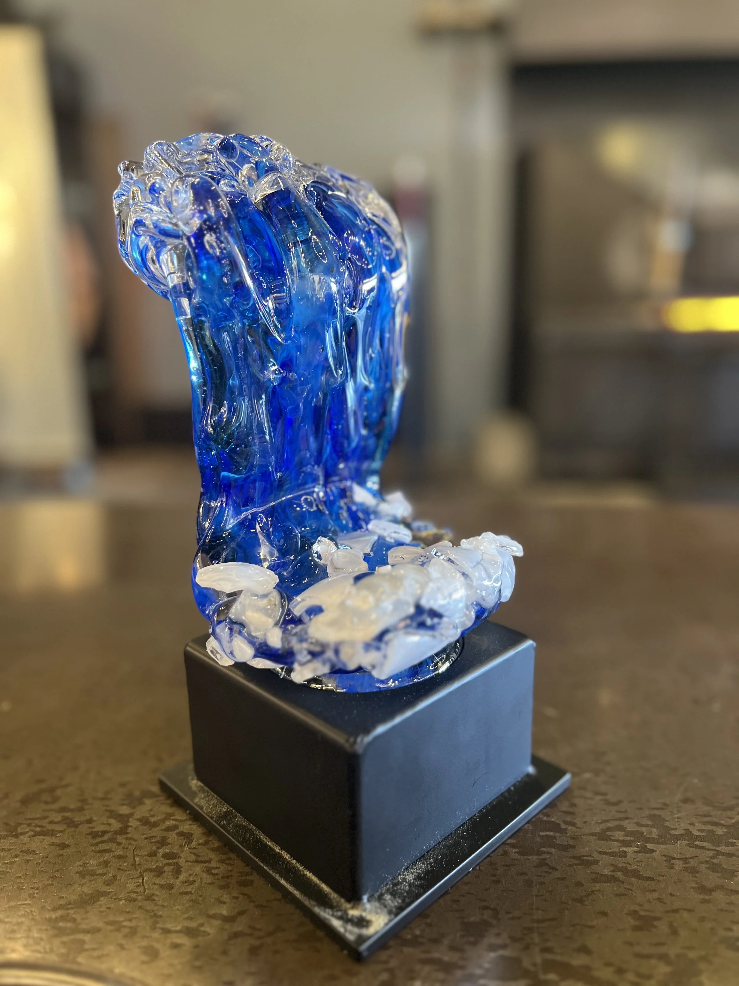 A glass sculpture featuring a blue wave design, mounted on a black base, placed on a brown surface.